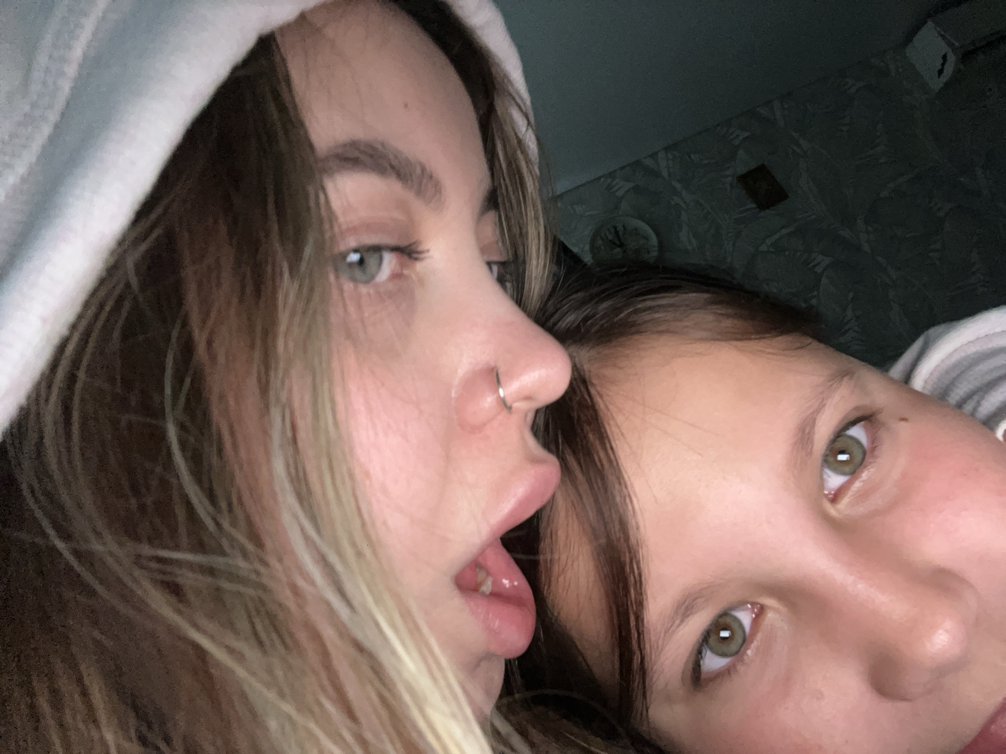 Two young women are positioned closely together, likely in an indoor setting. The woman on the left has light brown hair with blonde highlights and is wearing a white hooded sweatshirt. She has a nose piercing and her mouth is slightly open. The woman on the right has dark brown hair. Both women have fair skin and light-colored eyes. The background is dark and blurred, with a patterned wall visible in the upper right corner. It is unclear if they are located in Novopokrovskaya, Russia.