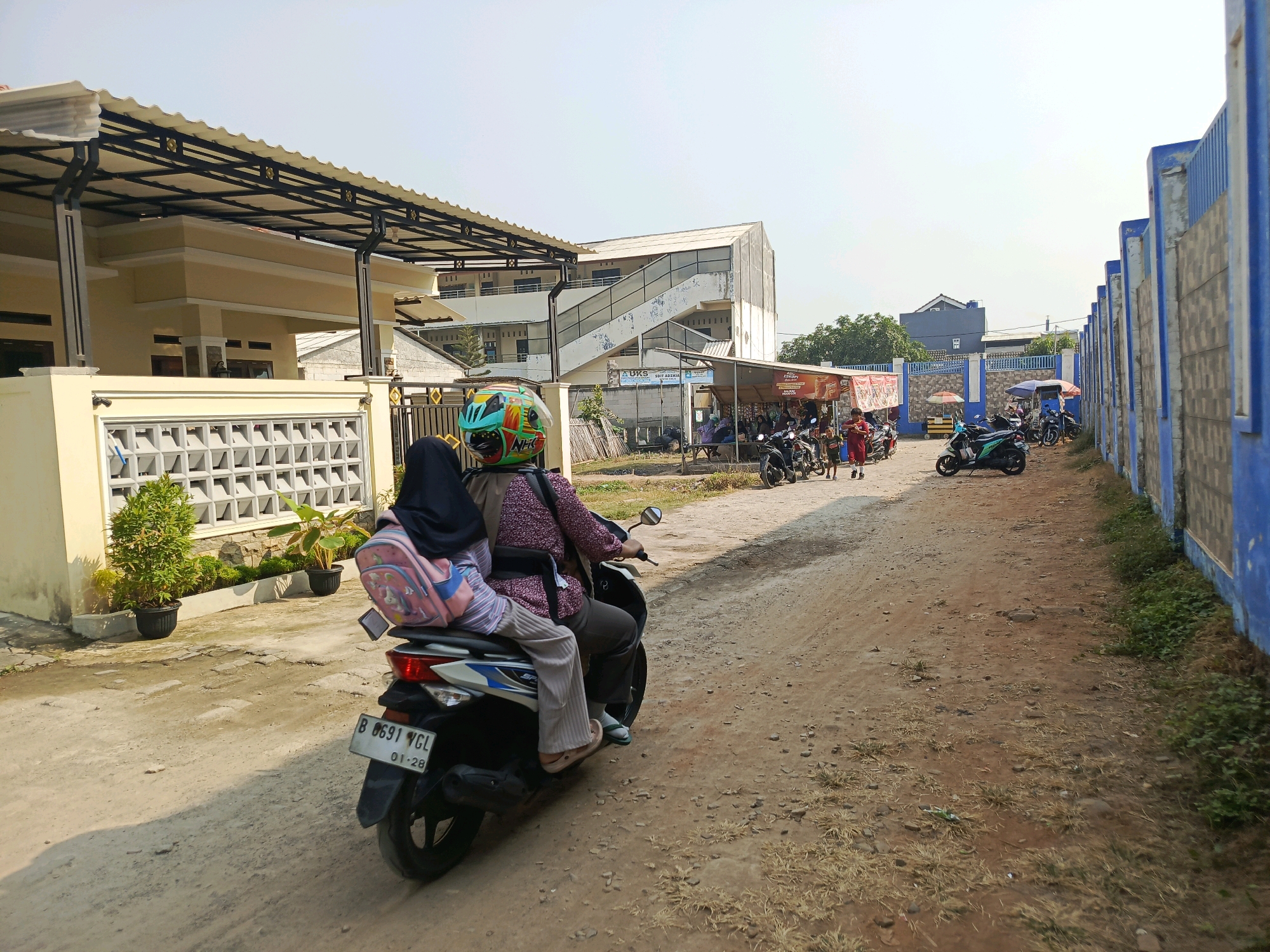 This image depicts a dusty, unpaved street scene in the city of Jati Mulya, Indonesia. In the foreground, a motorcycle carries two individuals. The rider, wearing a helmet with colorful decals and a patterned shirt, is operating the vehicle. The passenger, seated behind the rider, wears a black hijab and a light-colored long-sleeved shirt, with a pink backpack adorned with cartoon characters secured to their back. The motorcycle's license plate reads "B 6691 VGL". To the left of the street, a beige building with a white-roofed overhang is visible. This building features decorative wall vents and potted plants. Further down the street on the left, a multi-story building with an external staircase is present, and several small stalls or shops line the road. To the right of the street, a tall blue and grey concrete wall runs along the perimeter. Several other motorcycles are parked along the street and near the stalls. The overall environment is characterized by dry, dusty ground, sparse vegetation, and a clear, bright sky. The atmosphere suggests a typical day in a residential or commercial area of Jati Mulya.