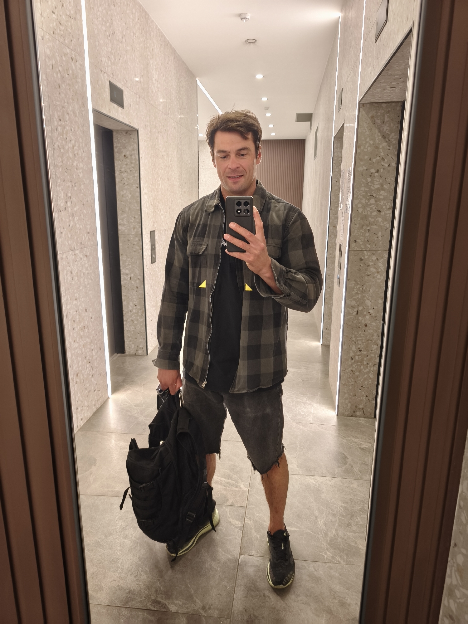 A man stands in a hallway, presumably indoors, holding a black backpack in his left hand and a smartphone in his right. He is dressed in a black t-shirt, a grey and black plaid jacket, and grey jean shorts, wearing black shoes. The hallway appears to have grey marble flooring and walls covered in a light-colored speckled pattern. Two elevator doors are visible on the left side of the frame. Thin, vertical lights run along the edges of the elevator bays. The man seems to be taking a picture of himself in the mirror. The reflections of the hallway suggest this is a modern building. The location is likely in the city of Moscow, Russia.