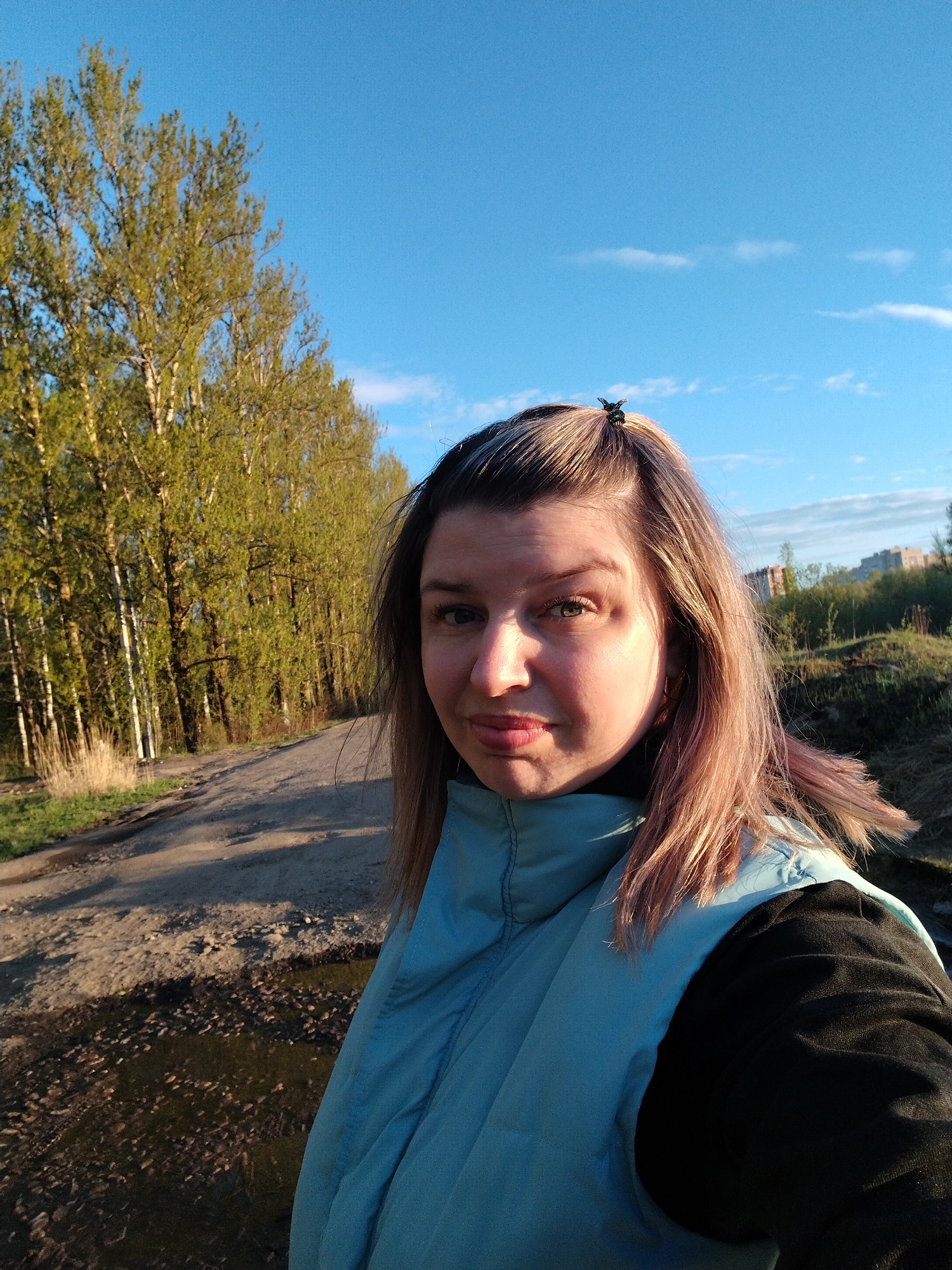A woman is posing for a selfie on a dirt road. The location is likely Fedorino, Russia. She is wearing a light blue vest over a black long-sleeved shirt. Behind her is a line of trees and a blue sky with a few clouds. The dirt road has puddles. Buildings are visible in the distance.
