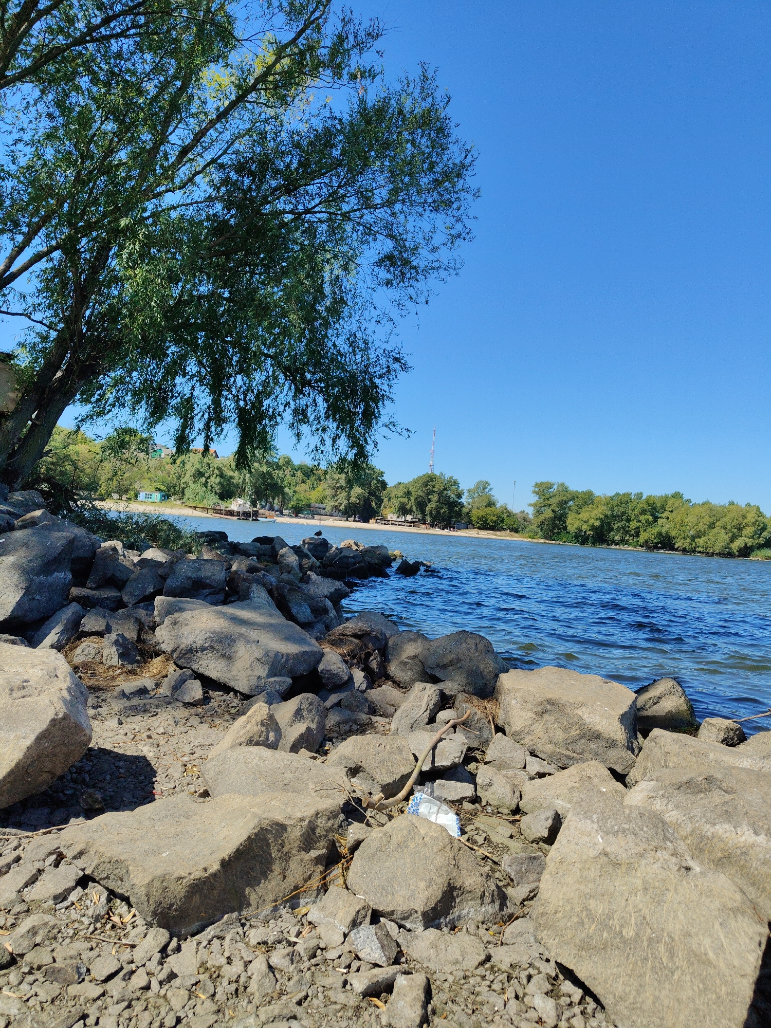 The image depicts a rocky shoreline of a river on a clear, sunny day. The foreground is dominated by large, gray stones and smaller gravel. A single white plastic bag lies among the rocks. A large tree with green foliage overhangs the left side of the frame. In the midground, the blue water of the river is visible with gentle ripples. The opposite bank is lined with dense green trees, and in the distance, there are some buildings and a tall radio tower. The overall scene suggests a natural landscape within a city, possibly a park or recreational area along the Dnieper River in Cherkasy, Ukraine. There are no people visible in the image.