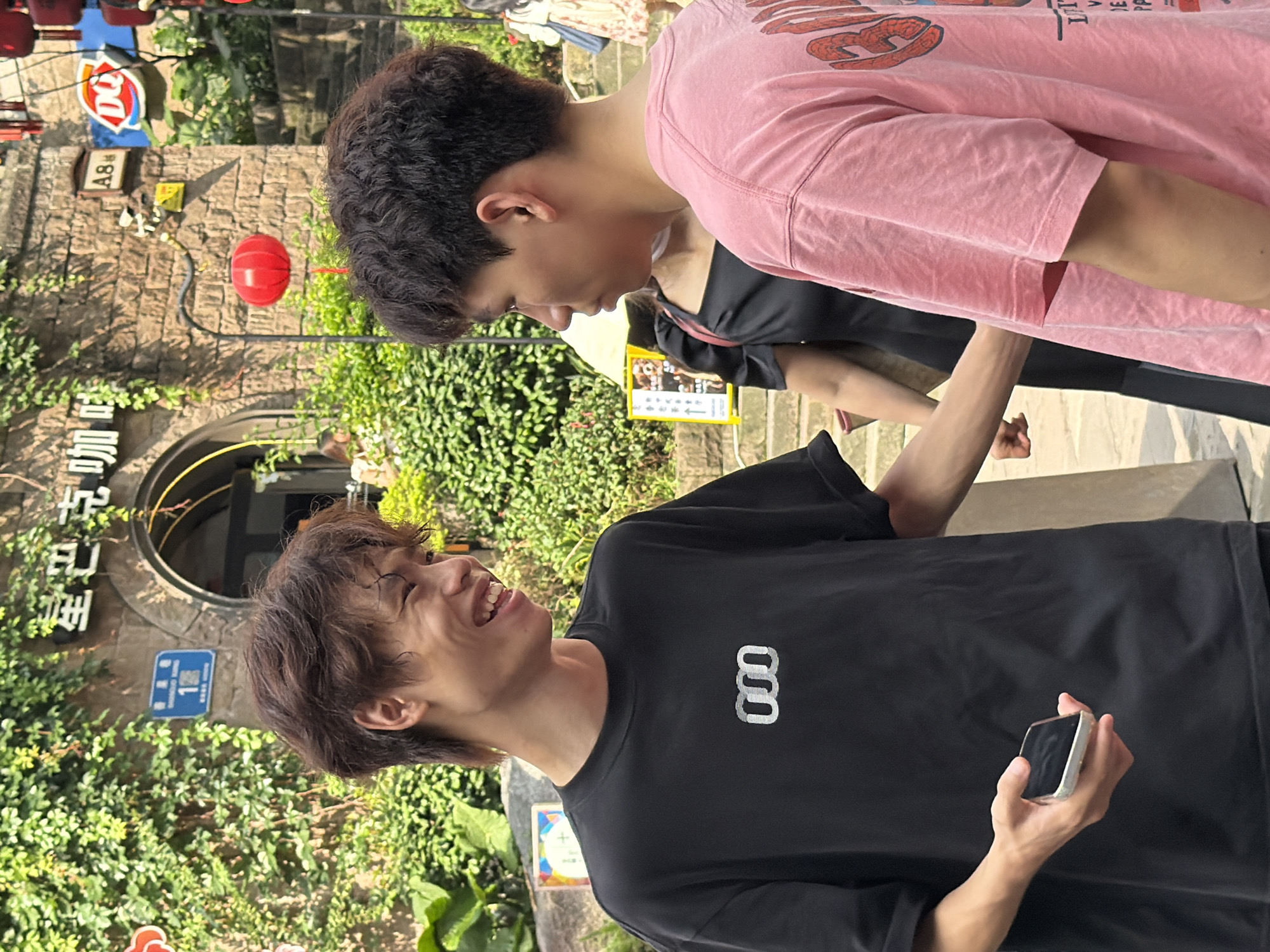 Two young males, appearing to be in their late teens or early twenties, are the central figures in this outdoor scene. The male on the left, wearing a black t-shirt with a white abstract logo and holding a smartphone in his left hand, is smiling broadly and looking towards the male on the right. The male on the right, facing away from the camera and wearing a pink t-shirt with graphics on the back, is looking towards the male on the left. They appear to be engaged in conversation.
The background suggests an urban environment, possibly a street or pedestrian area in Yuzhong District, China, given the presence of Chinese characters on signage and architectural elements characteristic of such locations. A building with a stone facade and an arched entrance is visible behind the individuals. Lush green foliage covers parts of the building and the surrounding area, indicating a pleasant, possibly warm weather day. A red lantern hangs from an overhead structure. Other people are partially visible in the background, including a woman in a black dress and another person in a floral dress. The overall atmosphere is casual and social.