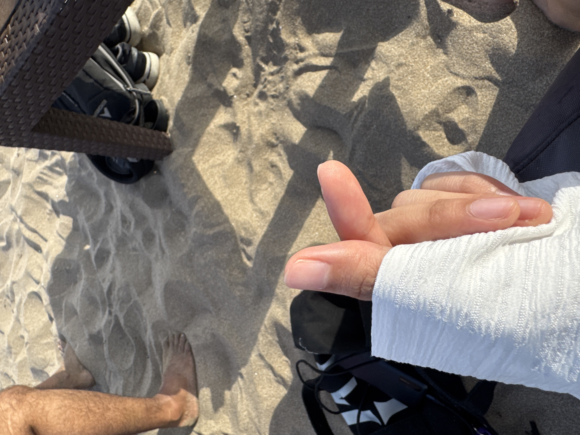 A first-person perspective shot shows a hand forming a Korean heart gesture with thumb and index finger, wearing a white textured long-sleeved shirt, placed on a sandy beach. In the background, a person's bare legs and feet are visible, along with a dark bag and a pair of sneakers partially hidden under a wicker chair. The sandy ground casts shadows from the chair, suggesting it is daytime. The scene is set on Kuta Beach in Kuta, Indonesia.