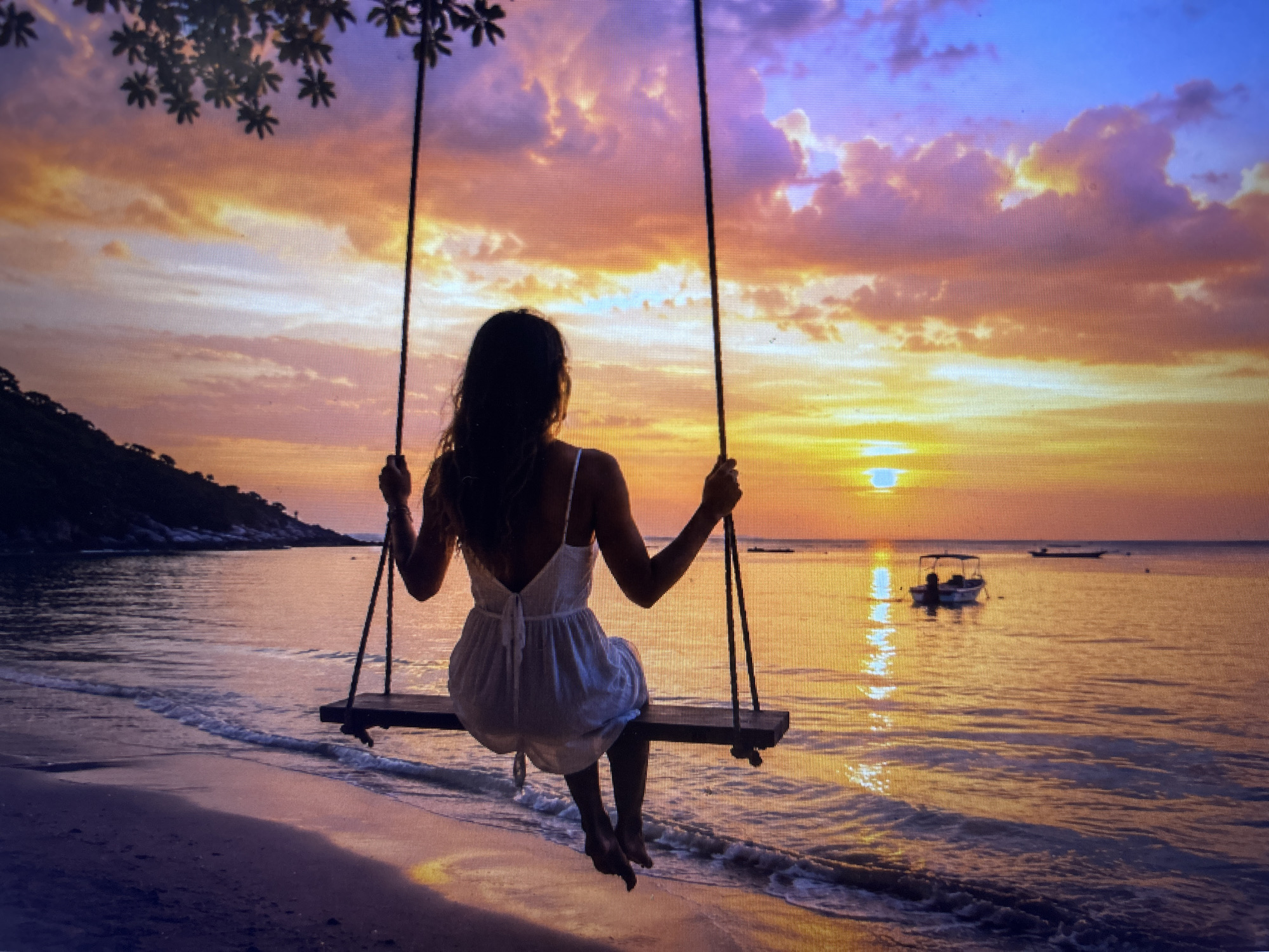 A woman in a white dress sits on a swing at a beach as the sun sets. The sky is a mix of orange, yellow, purple, and blue hues. Boats are visible on the water in the background. Trees are seen on the left side of the image.
There is no evidence the location is Sofia, Bulgaria.