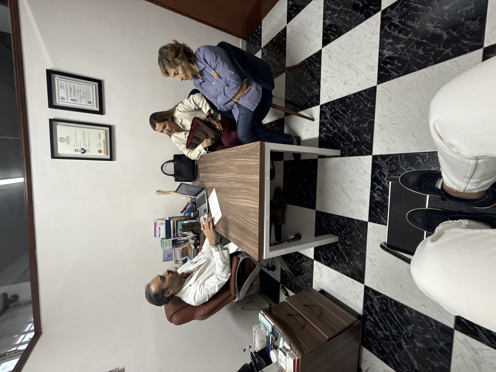 The scene depicts a doctor's office, likely in Puebla, Mexico, based on the framed certificate bearing the name "Universidad de Oriente." The office has a black and white checkered floor and white walls adorned with two framed certificates. A man in a white doctor's coat sits at a wooden desk, reviewing a paper. Two women stand beside the desk; one older woman in a blue striped shirt appears to be clutching her stomach, while the other woman in a white jacket leans over the desk, appearing to consult with the doctor. Various items clutter the desk, including a laptop, books, and other documents. A small wooden cabinet is adjacent to the desk. The lighting is bright, suggesting a daytime setting.