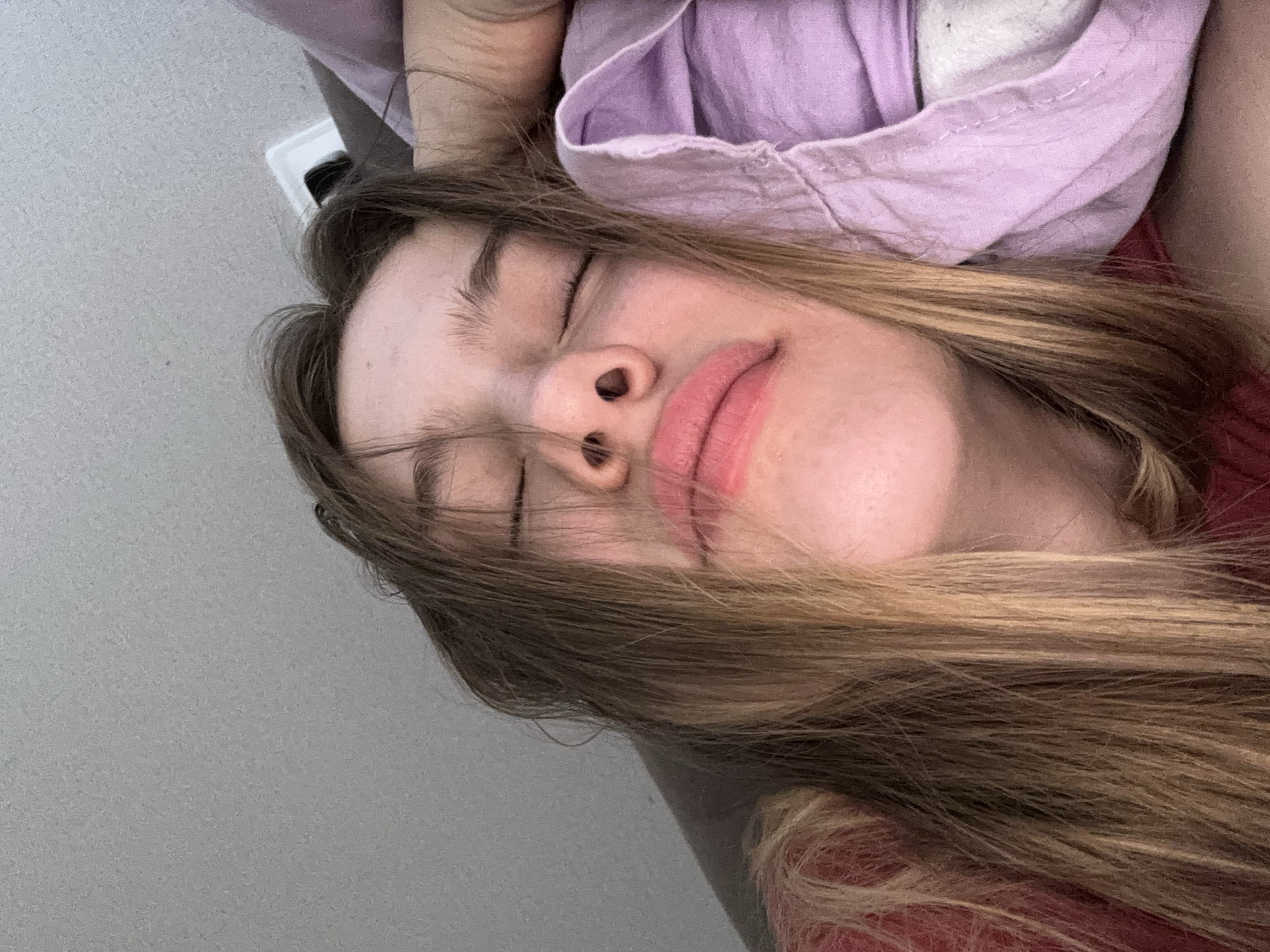 A young female with light brown hair is lying down, her eyes closed. She is wearing a pink top and is resting her head on a pillow with a light purple cover. The background appears to be a wall. The image was taken indoors in Vsevolozhsk, Russia.