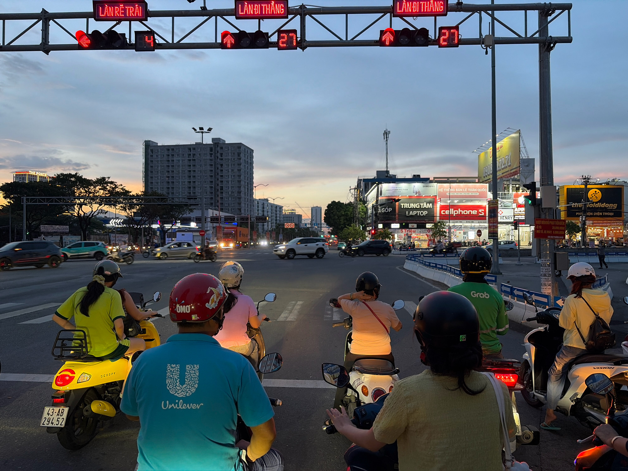 The image depicts an urban street scene in Đà Nẵng, Vietnam, at dusk. A traffic light overhead displays red signals and the numbers "4" and "27". Several people on motorbikes are stopped at the intersection, waiting for the light to change. The rider in the foreground on the left wears a blue shirt with a Unilever logo and a red helmet. To their right, another rider wears a black helmet and a light-colored shirt. In the middle ground, a rider in a yellow shirt is on a yellow scooter, and behind them, another rider in a pink shirt. Further back, a rider in a green jacket with a "Grab" logo is visible. Buildings and billboards line the street in the background, indicating commercial activity. The sky is filled with twilight colors.