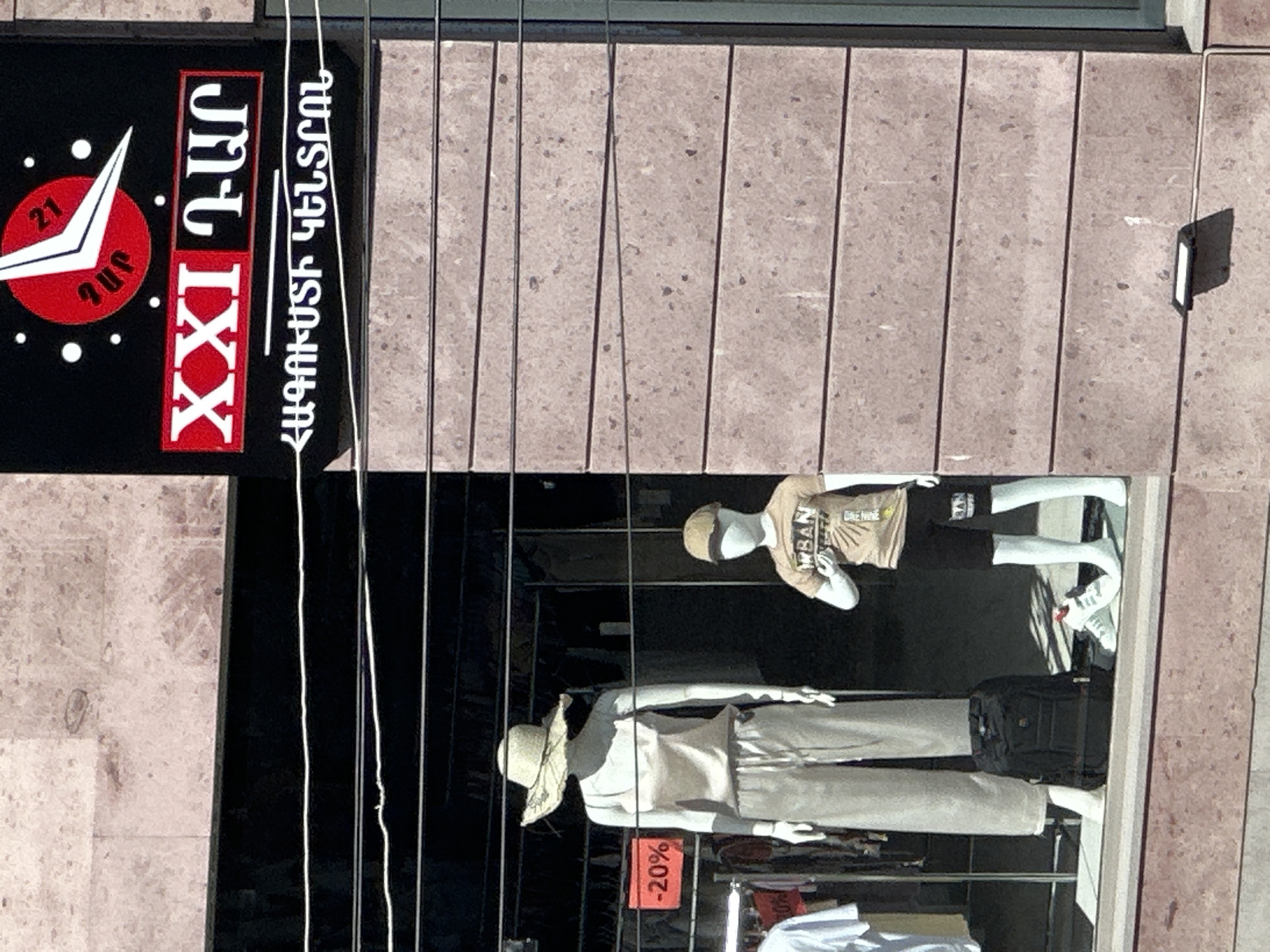 The image shows a street-level storefront in Gyumri, Armenia. A sign above the storefront reads "XXI ԴԱՐ" and has a clock graphic. Below the sign, in Armenian script, is additional text. In the storefront window, there are two mannequins. The left mannequin is wearing a light-colored tank top and matching wide-leg pants, with a straw hat. A black backpack is positioned at its feet. To the right of this mannequin is a smaller mannequin dressed in a tan t-shirt with the word "URBAN" on it, and black shorts, wearing a tan baseball cap. A pair of white sneakers is placed in front of the smaller mannequin. Red sale tags displaying "-20%" and "0%" are visible on racks of clothing in the background. The building exterior is constructed of pinkish-brown stone blocks. Wires are visible across the storefront window.