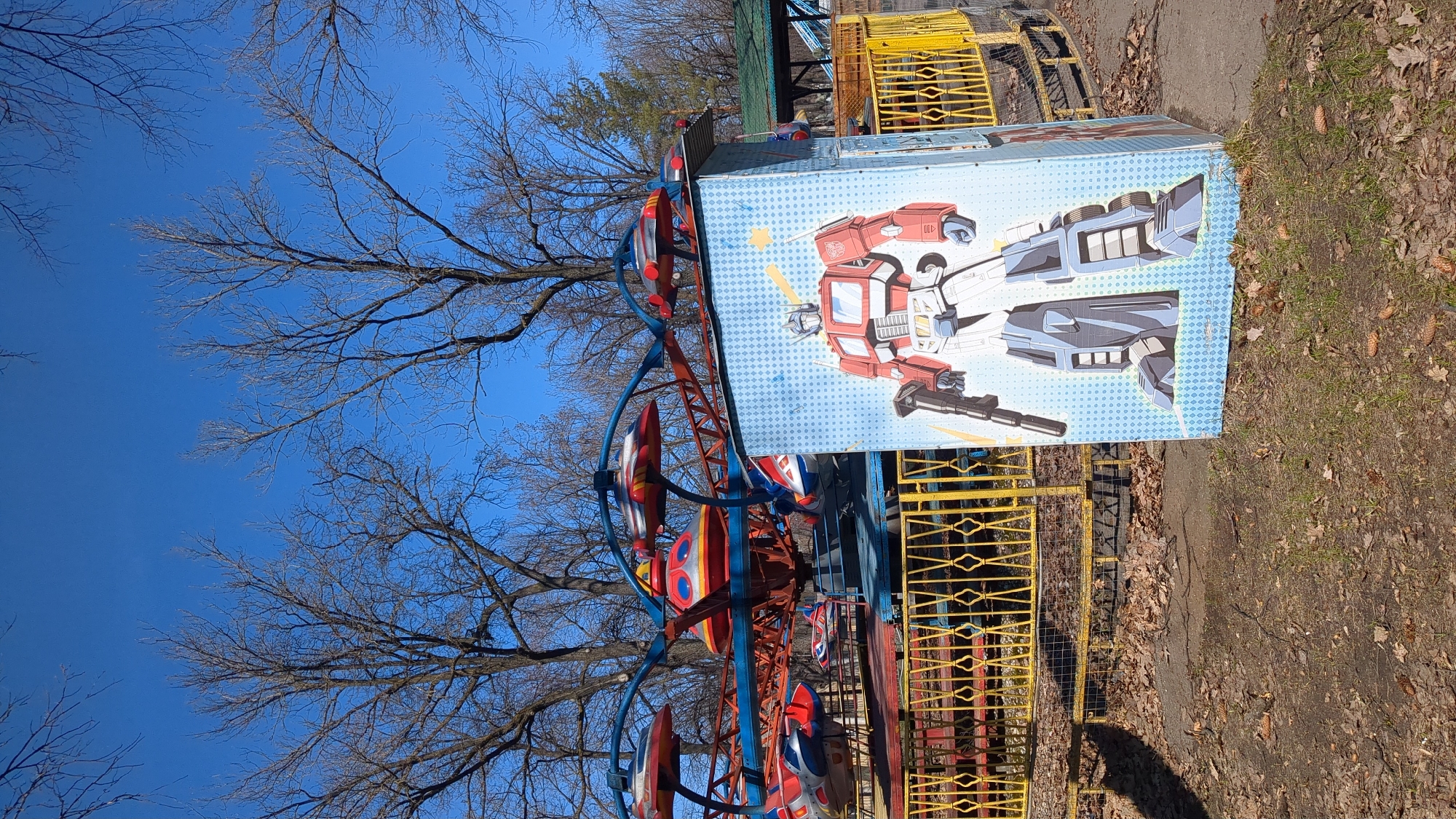 In Ulyanovsk, Russia, a colorful amusement ride stands in a park, featuring red, white, and blue rocket-shaped cars suspended from a blue metal frame. A large, rectangular sign with a drawing of a transformer is attached to the side of the ride. A yellow metal fence surrounds the ride, and the ground is covered with dry leaves. Bare tree branches reach towards a clear blue sky in the background.