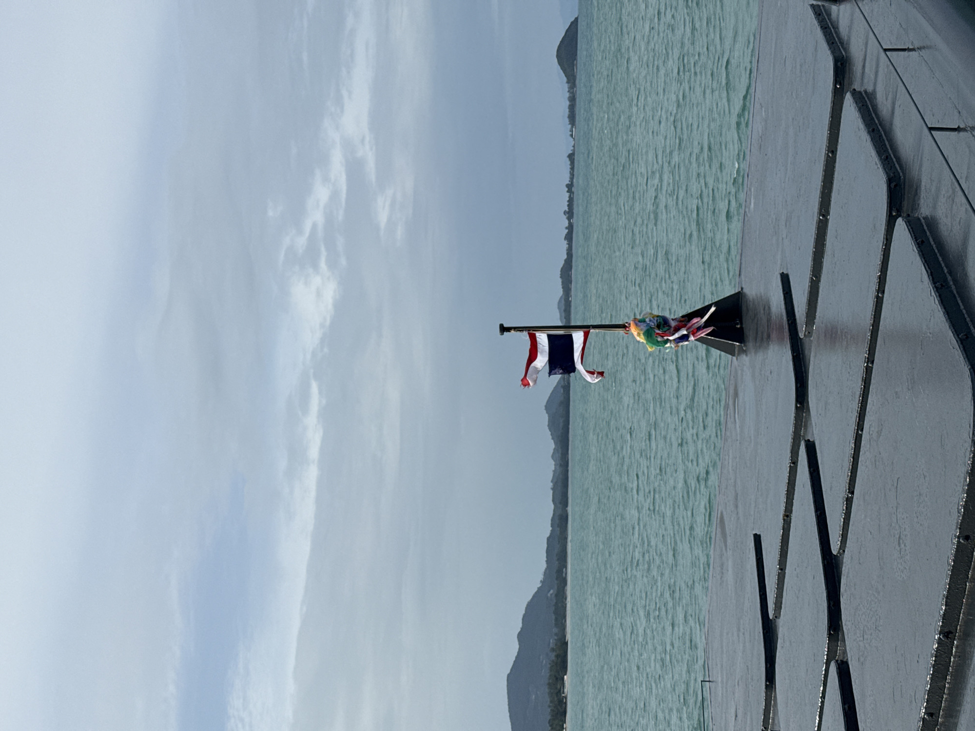 The image captures a view from what appears to be a ferry or boat in the Ko Samui District of Thailand. In the foreground, a dark, wet surface composed of several rectangular panels with rounded corners occupies the bottom portion of the frame. A flagpole stands prominently on this surface, flying the Thai flag. Colorful prayer flags or ribbons are tied around the base of the flagpole. The sea, a vibrant turquoise, stretches out to the horizon, with gentle ripples on its surface. In the distance, a coastline with lush green hills and trees is visible. The sky is overcast with scattered clouds, suggesting a potentially cloudy day. The overall scene is serene and depicts a typical coastal landscape in Thailand.