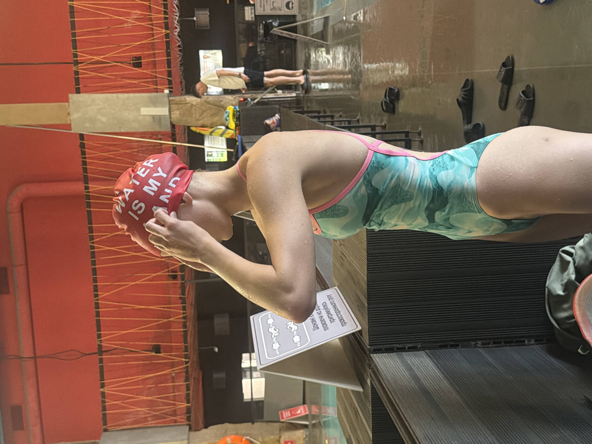 The image depicts a person wearing a red swimming cap with the words "WATER IS MY LAND" in white lettering, and a teal and pink swimsuit. The person is standing poolside and adjusting their swimming cap. The setting appears to be an indoor swimming pool with red walls and orange netting in the background. There is a sign with illustrations and text in a foreign language to the left of the person. Several pairs of black flip-flops are scattered on the wet floor. In the background, another person is visible standing near a railing. The overall environment suggests a public swimming facility. The location is identified as Kyiv, Ukraine.