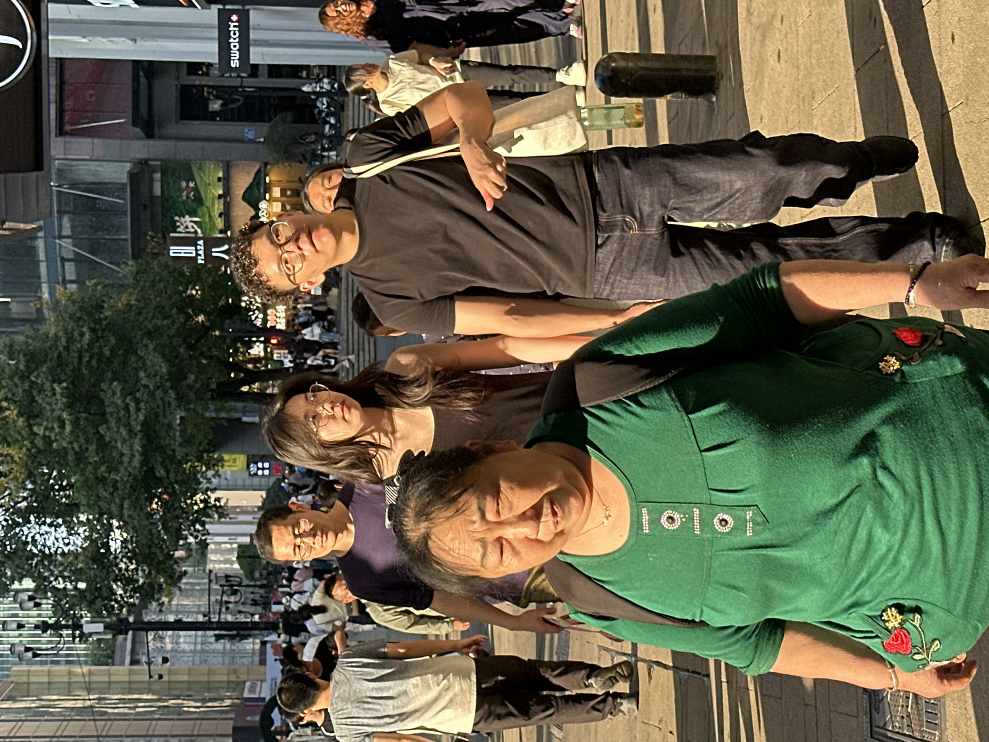 The image captures an outdoor urban street scene, likely in a bustling commercial area of Yuzhong District, China. The perspective is from within a crowd of pedestrians, with several individuals in the foreground and mid-ground looking towards the camera.
In the immediate foreground, an older woman is prominently featured, smiling warmly. She wears a vibrant green top adorned with decorative buttons down the placket and a red and yellow floral embroidery on her right sleeve. Her dark hair is pulled back, and she carries a dark bag over her shoulder.
Behind her and slightly to the right, a younger man with curly dark hair and glasses, dressed in a black T-shirt and blue jeans, also looks towards the viewer. He has a light-colored bag slung over his shoulder. Further back, a woman with long dark hair and glasses is visible, looking at the camera, next to a man in a dark purple shirt. Many other people are scattered throughout the background, indicating a busy environment.
The setting includes multi-story buildings, some with reflective glass facades, and a street lined with trees. The lighting suggests it is daytime, possibly late afternoon or early evening, given the long, distinct shadows cast on the pavement by the people and nearby structures. Two bottles, one dark and one light green, are standing on the ground to the right of the foreground figures.
Visible text includes the brand "Swatch+" displayed on a black rectangular sign attached to a building. On another building facade, the Chinese character "八" (bā) is seen above the English word "PLAZA". The scene depicts a lively moment of daily life in a contemporary Chinese city.