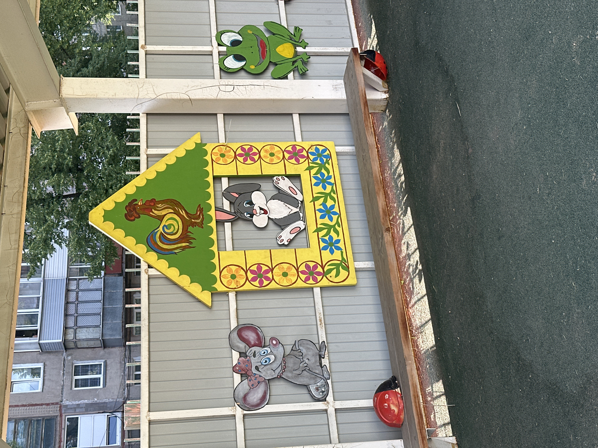 The image depicts an outdoor scene in Novoaltaysk, Russia, likely a playground or a similar public space. In the foreground, a weathered wooden bench runs horizontally. On the bench, to the left, is a red and black toy ladybug. To the right, another red and black toy ladybug is partially visible. The background features a grey corrugated metal fence decorated with colorful cartoon characters.
On the fence, from left to right, is a large, gray cartoon mouse wearing a pink bow and necklace. Next to the mouse is a yellow house-shaped cutout with green trim, adorned with floral patterns. Inside the window of the house, a gray and white cartoon rabbit is visible. To the right of the house is a green cartoon frog with large eyes and red lips.
Behind the fence, a building with multiple windows and balconies is visible, suggesting an urban environment. Trees with green foliage are also present in the background. The ground in the foreground is a dark, speckled surface, possibly asphalt or rubberized playground material. The lighting suggests it is daytime with sunlight casting shadows. There are no people visible in the scene.