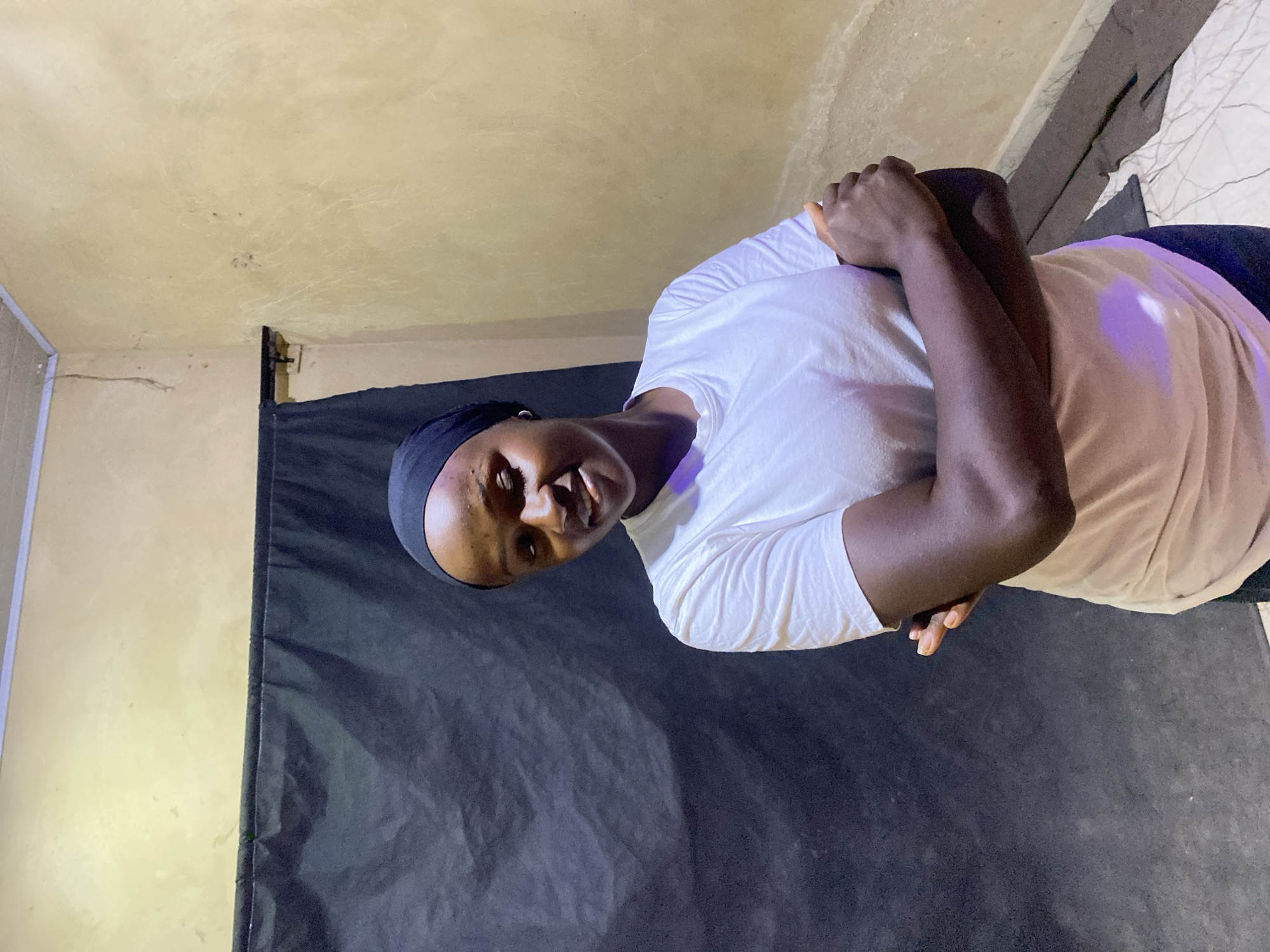 A young individual with dark skin, likely female, is captured in a half-body shot, looking directly at the viewer with a slight smile. She is wearing a plain white, short-sleeved t-shirt and a black headscarf that covers her hair. Her arms are crossed in front of her chest.
The setting appears to be indoors in Kuru Dungus, Nigeria. Behind the person is a dark, possibly black, fabric backdrop, which creates a plain background. Above the backdrop, a light beige or yellowish wall/ceiling with a textured surface and some faint marks or imperfections is visible. The lighting is somewhat dim, with a subtle purple hue reflecting on the white t-shirt, possibly from an artificial light source. The overall impression is a posed portrait in a simple, makeshift studio-like environment. There is no visible text in the image.