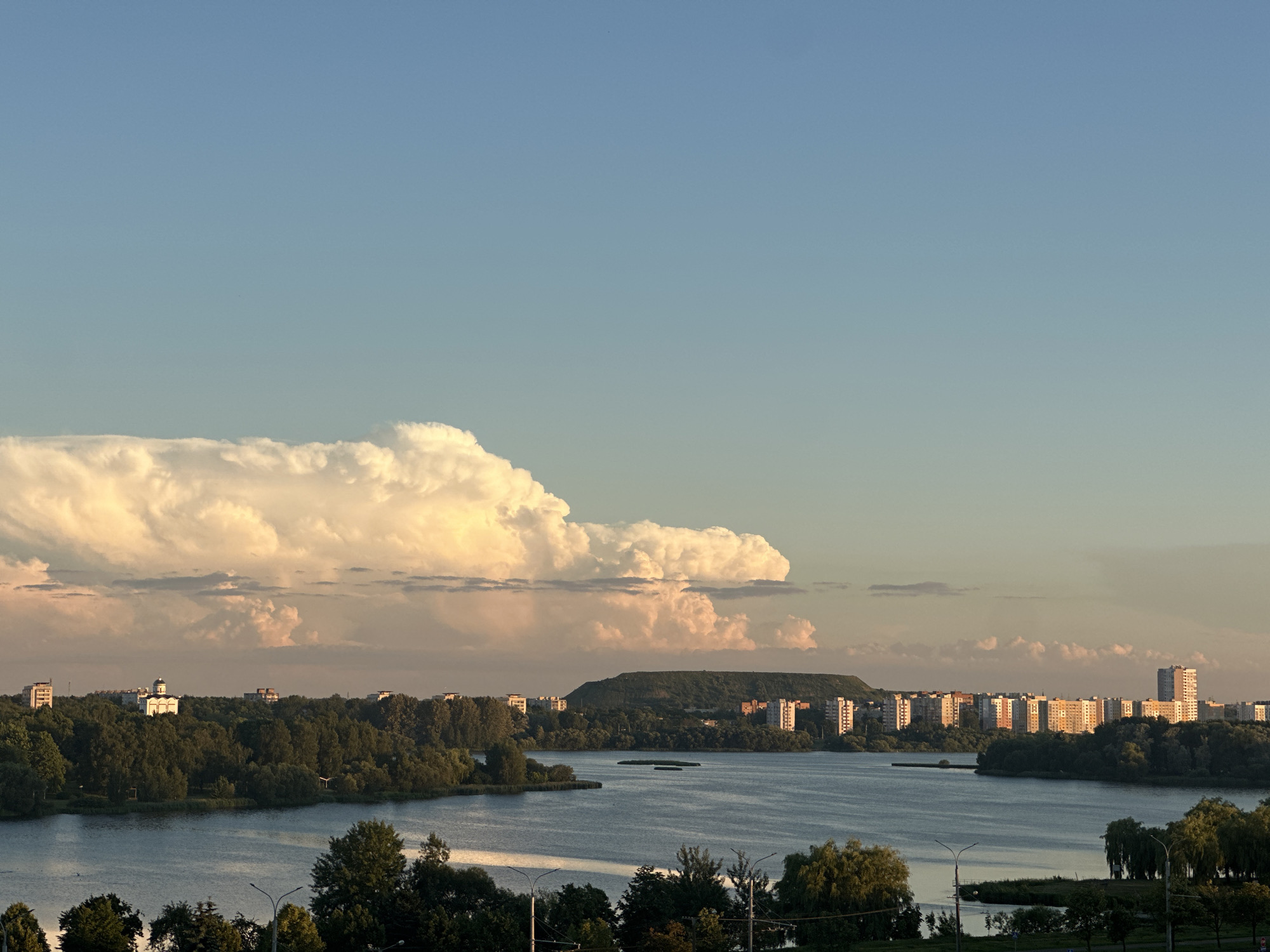 A wide shot captures a serene lake and its surrounding landscape in Minsk, Belarus, during what appears to be late afternoon or early evening. The sky is a clear, soft blue, gradually transitioning to a warmer, golden hue near the horizon. Dominating the upper left portion of the frame is a massive, cumulonimbus cloud formation, catching the golden light of the setting sun, its edges glowing with an almost ethereal warmth.
The lake itself is calm, reflecting the soft light and the muted colors of the sky and the distant shore. Along the far bank of the lake, a dense line of green trees forms a verdant barrier. Within this tree line, several buildings are visible, including apartment blocks with windows reflecting the sky's light. Most prominent among these is a white building with a dark dome, suggesting a church or religious structure, perched atop a slight rise. Further along the shore, a larger hill covered in vegetation rises, with more apartment buildings clustered at its base.
The foreground shows more trees and a glimpse of streetlights, indicating the proximity of urban infrastructure to this natural setting. The overall atmosphere is peaceful and tranquil, bathed in the warm, diffused light of the approaching sunset. No people or specific activities are clearly discernible in the image, emphasizing the quiet beauty of the landscape at this time of day.