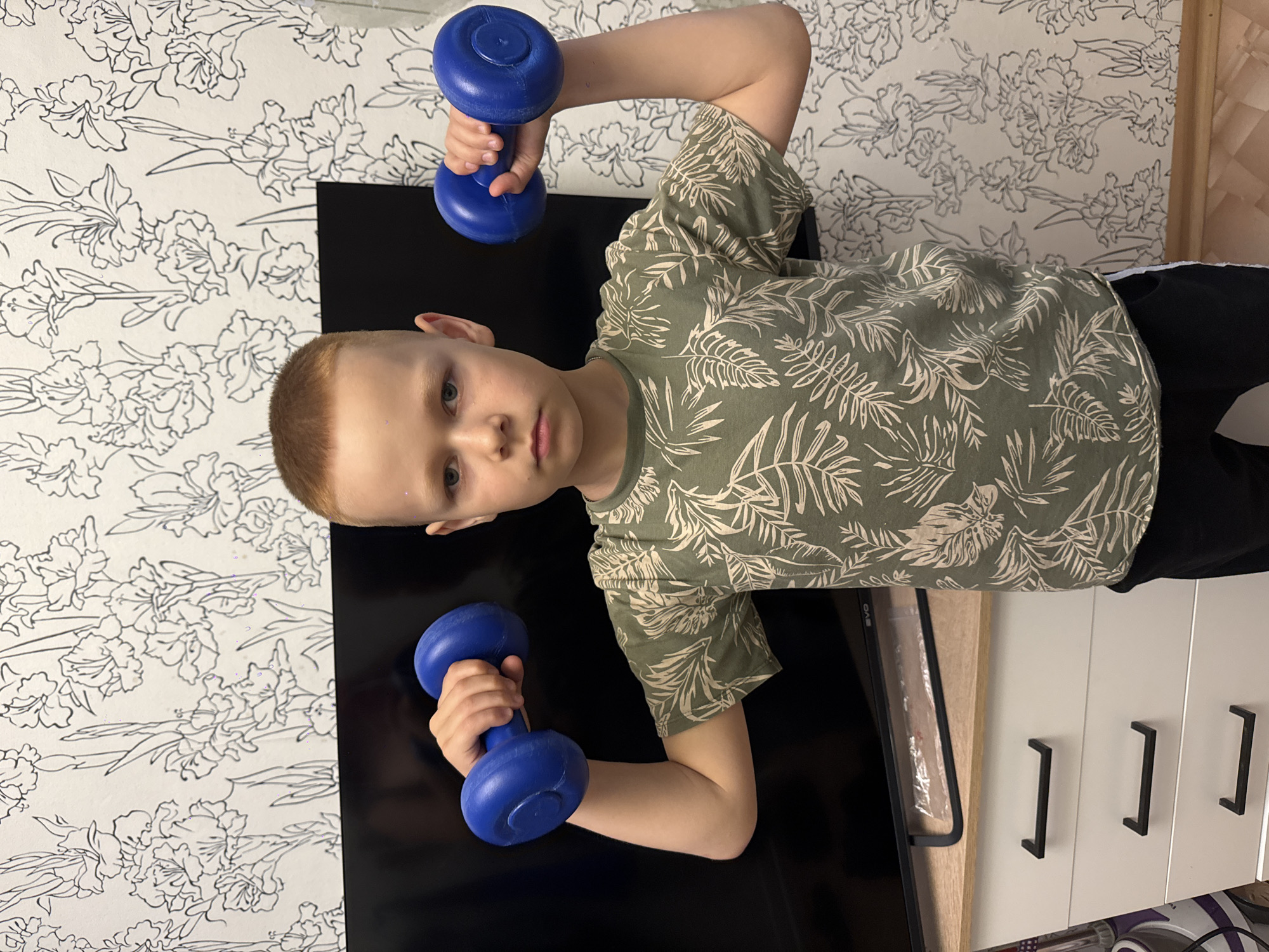 A young boy with blond hair and blue eyes is shown in a vertical, full-body portrait. He is wearing a green t-shirt with a palm leaf pattern. In his left hand, he holds a blue dumbbell, with his arm bent and elbow raised. In his right hand, he also holds a blue dumbbell, with his arm bent and elbow raised. The boy is looking towards the camera with a neutral expression.
The background consists of a wall with a floral wallpaper featuring black outlined flowers on a white background. To the right of the boy, a black surface, possibly a television screen, is visible. Below this, a white cabinet with three black handles is partially visible. The setting appears to be indoors. No specific time of day or weather conditions can be determined from the image. No visible text is present in the image. The overall impression is that of a child exercising at home.