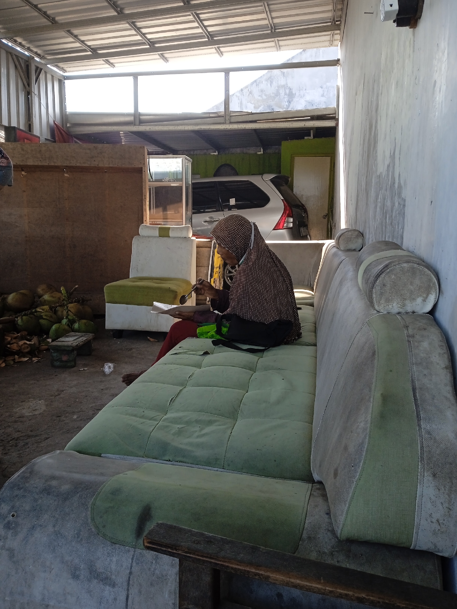 An elderly person wearing a brown-patterned hijab sits on a large, worn, green-and-gray sofa. The person is holding a spoon and a piece of paper, possibly eating or reading. Nearby are a pile of green coconuts and a small table. Behind them, there's a display case, a car, and a green door in a garage or carport setting. The location is in Brangsong, Indonesia, as indicated in the metadata. The overall setting appears to be a semi-outdoor space with a corrugated metal roof. The wall to the right is faded and has visible water stains. A light fixture is affixed near the top right of the image.