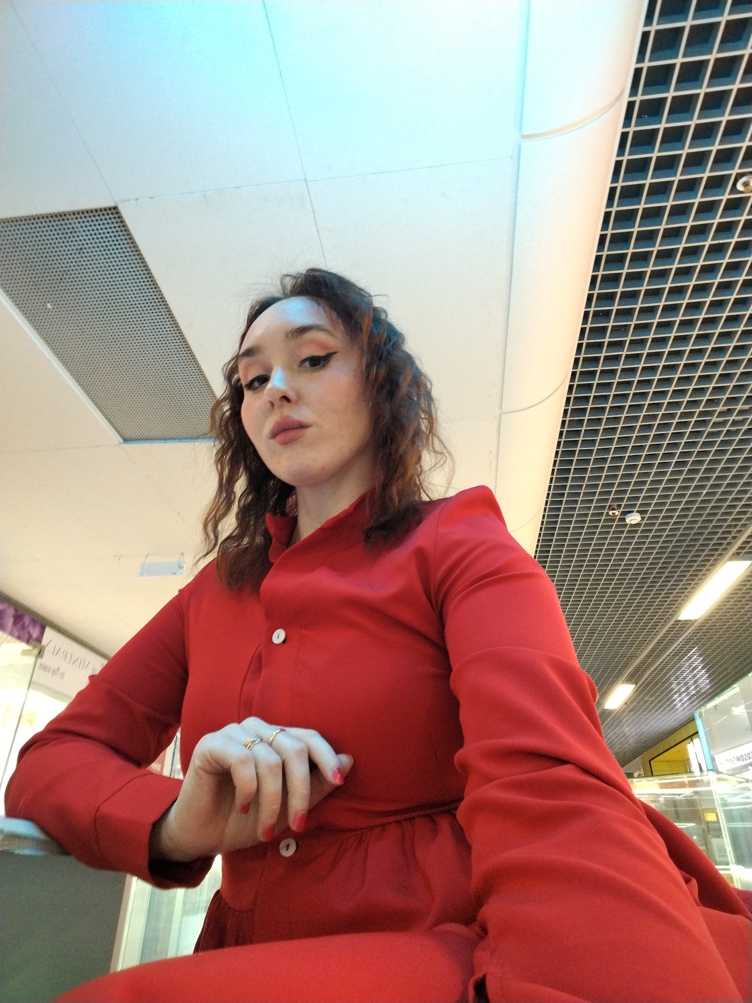 The media file shows a low-angle, indoor shot of a young woman with curly brown hair and fair skin. She is wearing a bright red dress with two white buttons down the front. Her makeup includes winged eyeliner and rosy blush. She is looking directly at the camera with a neutral expression. Her left hand rests on her lap, adorned with two gold rings and her fingernails are painted red.
The background features a modern interior with a white tiled ceiling and a grid-like structure above, illuminated by fluorescent lights. The lighting suggests it is daytime, but the specific time of day is not discernible. The environment appears to be a public space, possibly a shopping mall or a similar commercial building, given the glimpses of glass displays and signage in the background, some of which appears to be in Russian and contains legible text such as "IZHEVSK". The overall mood is calm and composed.