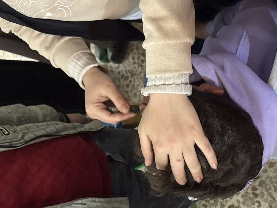 An overhead, close-up view depicts an indoor scene in Kafr Batna, Syria, involving at least three individuals. In the lower center, an individual with dark brown hair, presumed to be a child, has their head resting on a folded purple garment. A bare hand, originating from the right side of the frame and connected to an arm wearing a beige long-sleeved top with a ribbed cuff, rests gently on the top of the dark-haired individual's head. Another bare hand, also from the right and likely belonging to the same individual (indicated by the matching ribbed cuff higher on the arm), holds a small, blue and metallic dropper. This dropper is positioned above the dark-haired individual's facial area, appearing to administer a liquid. A third individual is partially visible on the upper left, wearing a grey zipped hoodie over a red shirt and dark pants. A bare hand from this third individual is partially discernible near the dropper. The floor is covered with light-colored, patterned tiles. The background beyond the subjects is dark and out of focus.