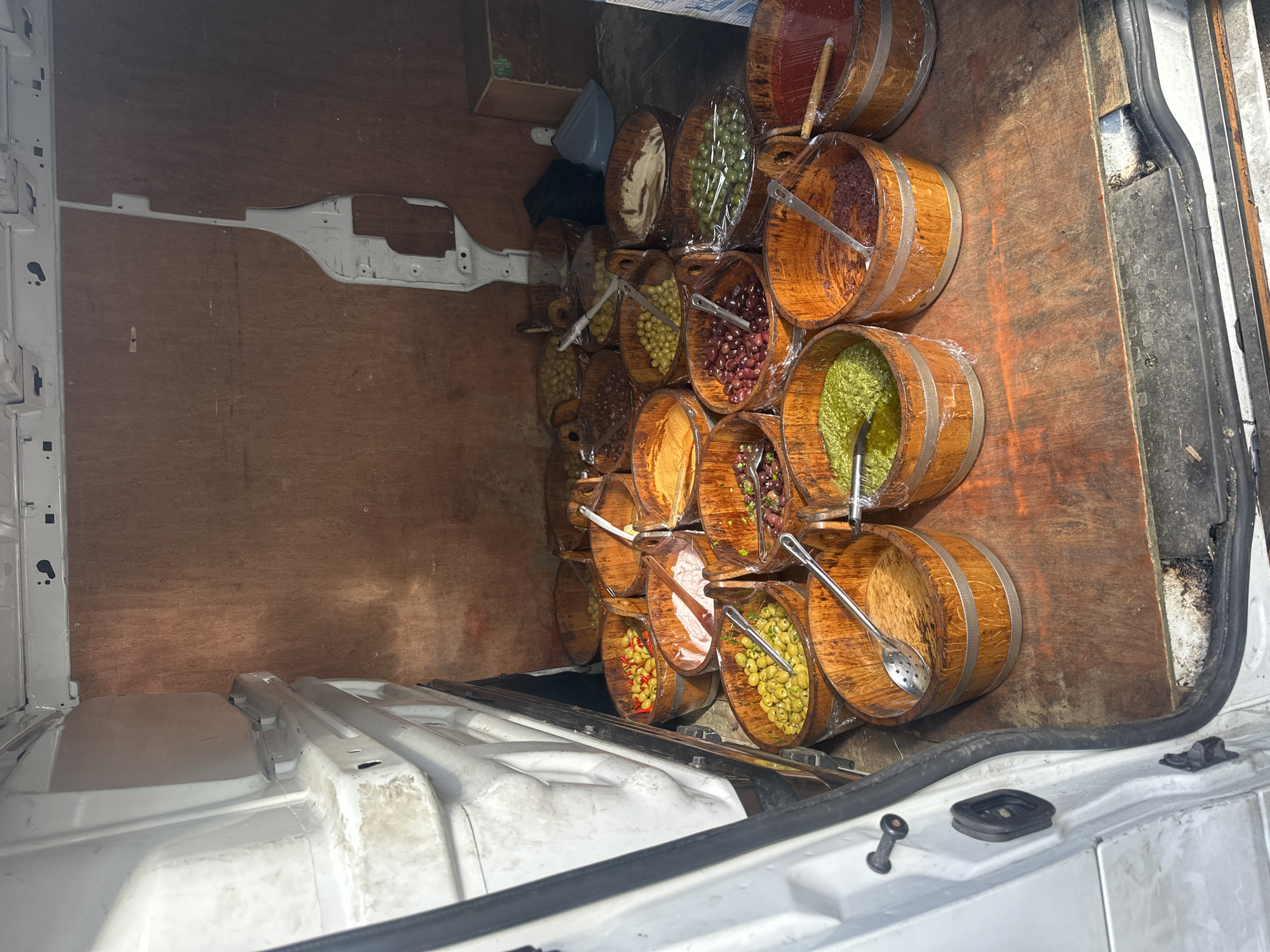 The image depicts the interior of a white van. A large number of wooden barrels, filled with what appear to be different types of olives and other preserved foods, are packed inside. Some barrels are covered with clear plastic wrap. A spoon rests in some of the barrels. The back of the van has wooden walls. The location is likely Sevenoaks, United Kingdom.