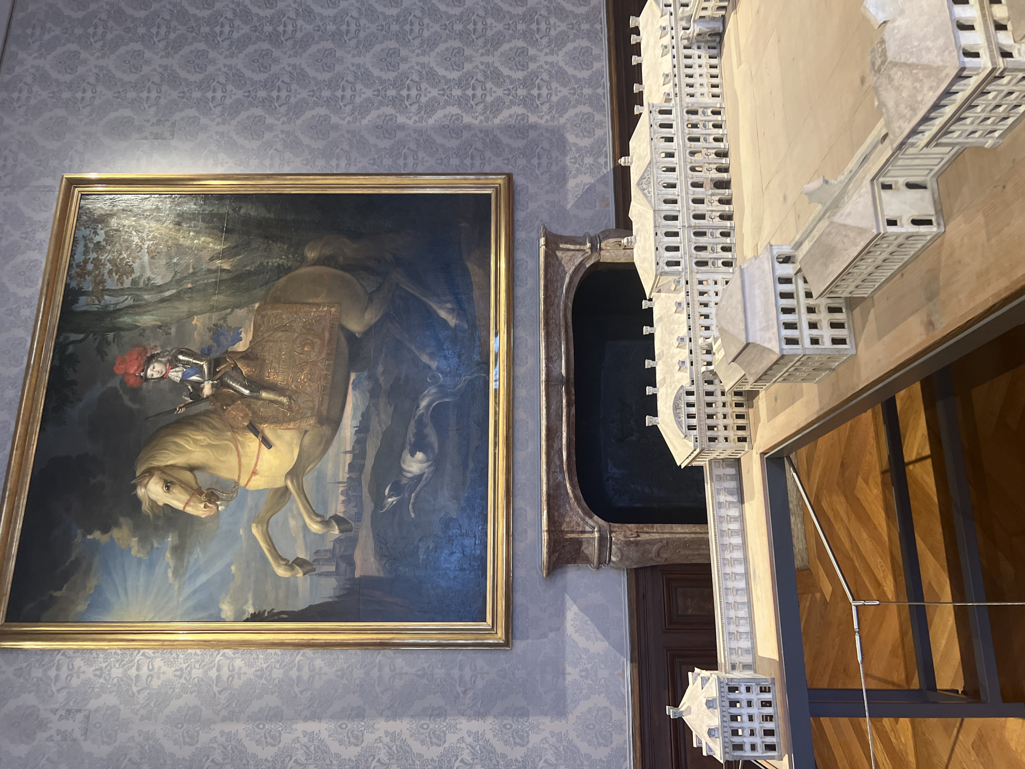 The image displays a room with ornate walls and a large painting above a fireplace. The painting depicts a young person in regal attire on a horse, with a dog running below. In the foreground, a detailed architectural model of a palace complex is laid out. The model includes multiple buildings with classical facades, courtyards, and connecting structures, representing a significant historical site. The room's ambiance suggests it is part of a museum or historical exhibition space, possibly within the Palace of Schleißheim in Oberschleißheim, Germany, given the scale and style of the model. The lighting is even, highlighting the details of the painting and the model. The walls have a damask pattern.