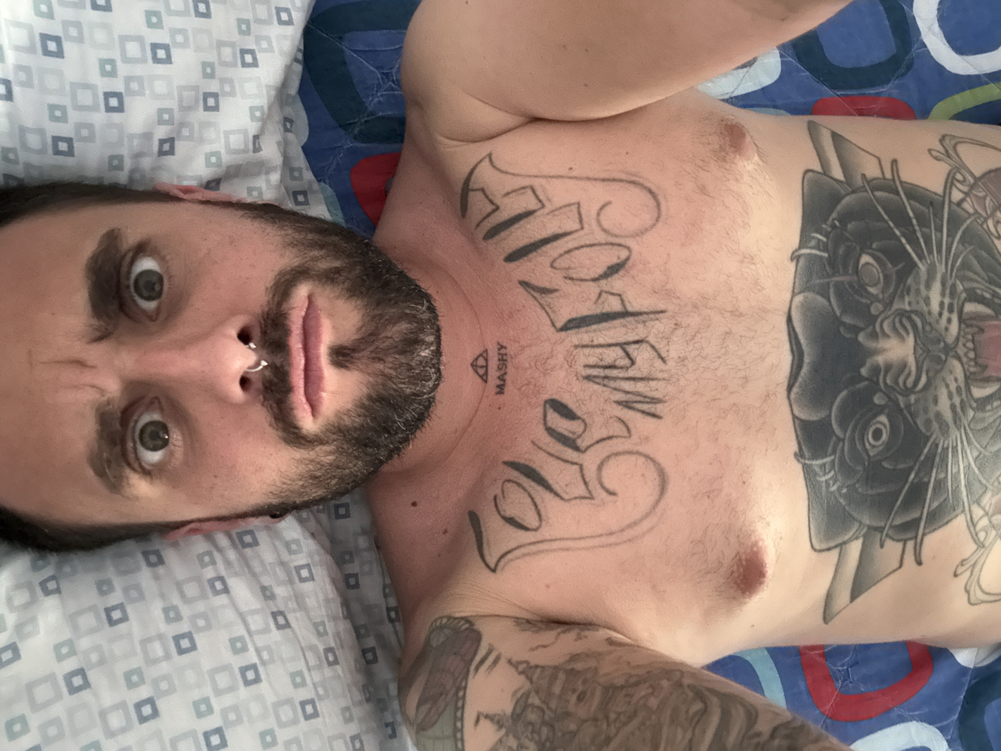 A man with a beard and a nose ring lies on a bed. He has tattoos on his chest and arm, including the words "FOREVER LOVE MY." The bedding is patterned with squares and circles. The location is unknown, and there is no indication of Aprilia, Italy.