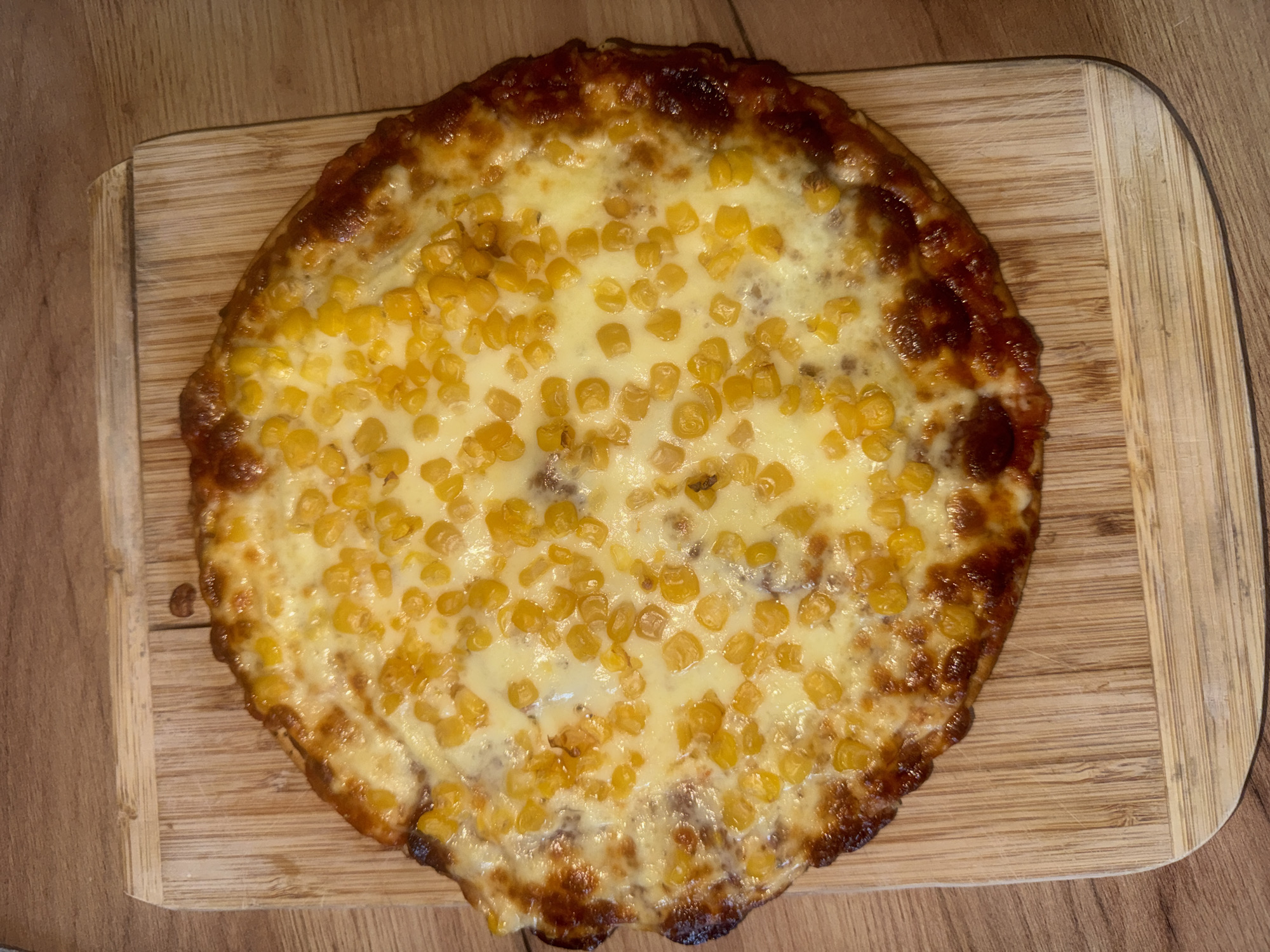 A close-up, overhead shot shows a round, golden-brown pizza topped with melted white cheese and scattered kernels of corn. The pizza has a slightly irregular crust, with some parts appearing darker and crispier than others. It sits on a light-colored wooden cutting board, which is placed on a darker wood surface. The lighting suggests an indoor setting, possibly a kitchen or dining area. No people are visible in the image, and the focus is entirely on the food. The scene is static, with no action depicted. The image is a still photograph, with no visible text. The location is identified as Varna, Bulgaria.