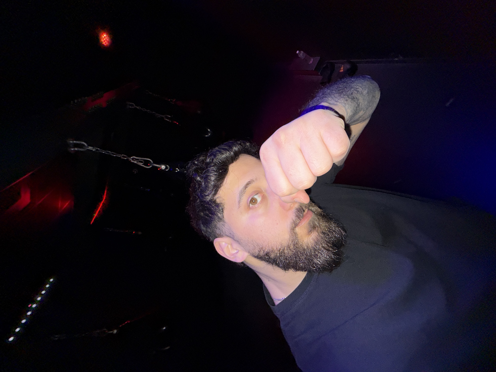 The scene is an interior view, likely a nightclub or bar, located within the city of Strasbourg, France.
A single male individual is depicted in the foreground, positioned centrally and facing the viewer. He possesses dark, curly hair and a dark, full beard. He wears a black short-sleeved t-shirt and a thin, silver-colored chain around his neck. His visible right eye is brown. His left forearm displays dark tattoo work, and he wears a dark band on his left wrist. His left hand is clenched into a fist and raised, partially obscuring the lower left portion of his face, specifically the area around his nose and upper lip.
The background is predominantly dark, illuminated by various artificial light sources. Multiple metal chains hang vertically from the obscured ceiling, some exhibiting visible snap hooks at their ends. Overhead, a linear arrangement of small, bright white circular lights is visible in the upper left quadrant. To the upper right, a diffuse red glow is present, accompanied by a distinct cluster of red LEDs and a horizontal red light streak. A blue light casts a glow upon the lower portion of the subject's shirt and the immediate foreground, indicating a light source from below or the left side of the frame.