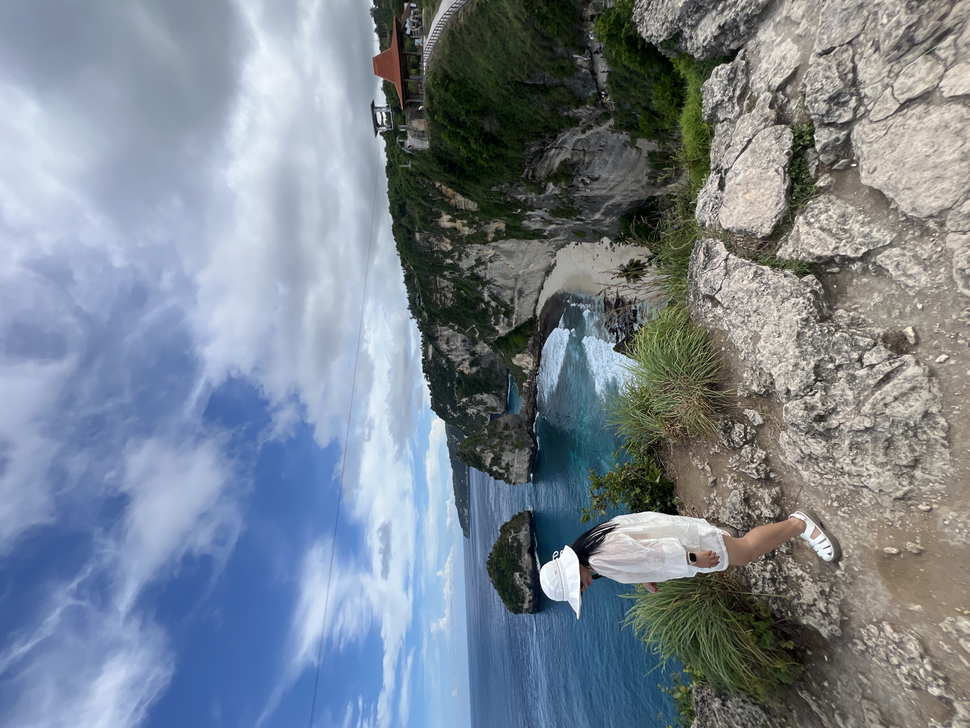 The image captures a person standing on a rugged clifftop overlooking a dramatic coastal landscape in Nusa Penida, Indonesia.
In the foreground, a person, likely female, is seen from behind and the side, standing on a rocky, uneven path fringed with green grass. She is wearing a white long-sleeved shirt, white shorts, white sandals, and a wide-brimmed white sun hat. An Apple Watch is visible on her left wrist. Her pose suggests she is observing the panoramic view.
The midground and background reveal a stunning seascape. Steep, vegetated limestone cliffs plunge into the ocean, with white waves breaking against their bases and a secluded sandy beach visible below, complete with palm trees. Several small, lushly vegetated islands and unique rock formations dot the turquoise and deep blue water, including one with a natural arch. On a higher part of the main cliff in the distance, a structure with an orange-tiled roof, possibly a viewpoint or resort, and what appears to be a lift or cable car mechanism are visible, along with a winding path. The sky above is a blend of clear blue with scattered white clouds, and some heavier grey clouds. The bright lighting indicates it is daytime. No text is visible in the image.