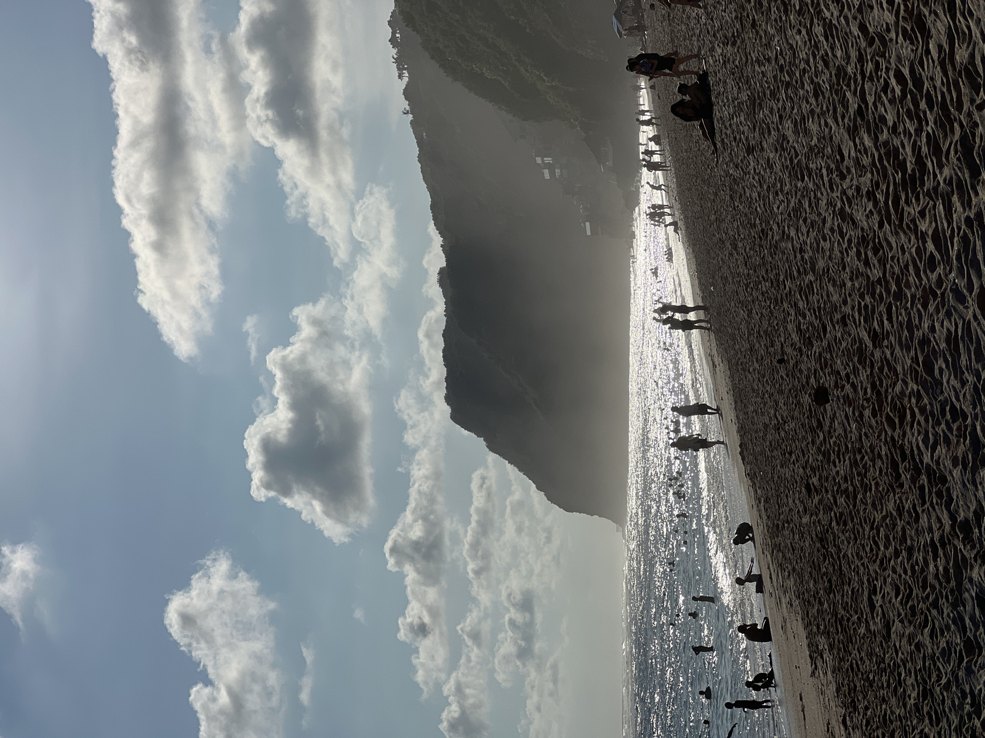 Ungasan, Indonesia: A beach scene shows a dark mountain against a partly cloudy sky. The sun reflects off the water as people stand and walk along the shore. The sand is a dark, coarse texture.