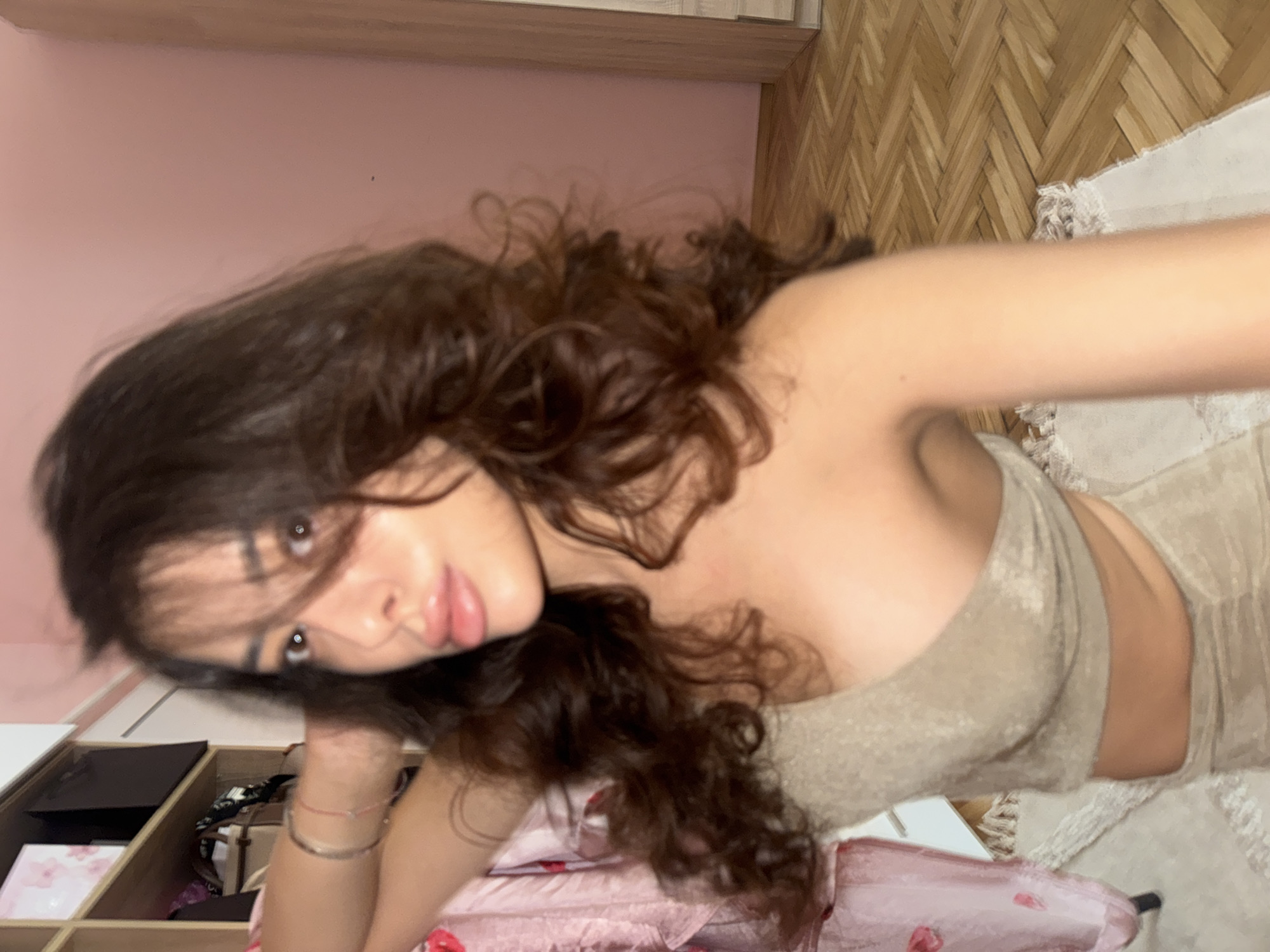 A young woman with long, dark, wavy hair is posing for a selfie in an indoor setting. She is wearing a beige, one-shoulder crop top and a matching skirt. Her left arm is raised, with her hand resting on the back of her head. Her right arm is extended, holding the phone to take the picture. The background shows a closet with shelves, a pink wall, and a wooden floor with a herringbone pattern. A white rug is visible on the floor in the foreground. The room appears to be a bedroom or dressing area. There is no information in the image to suggest the location is in Plovdiv, Bulgaria.