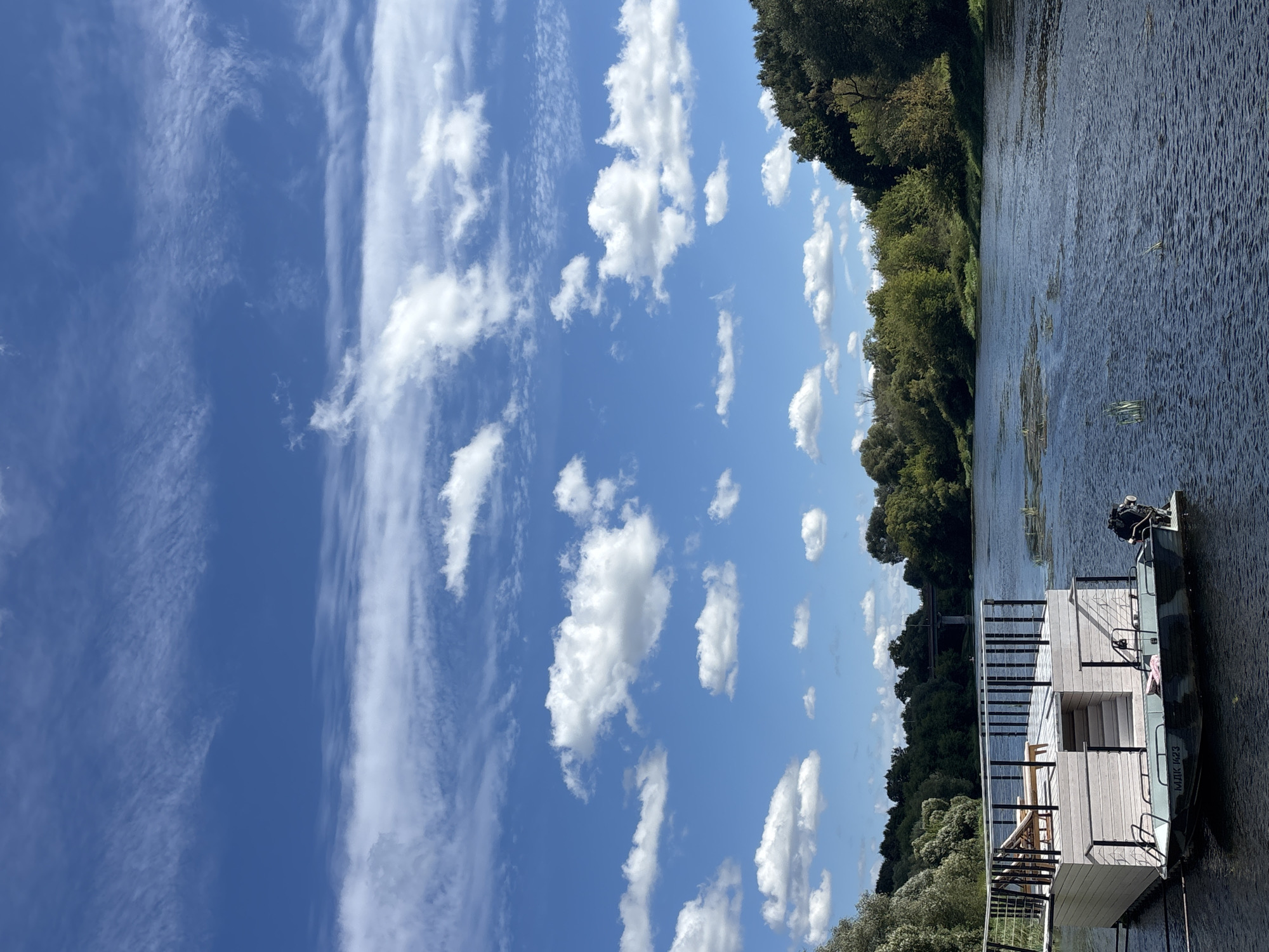 The image captures a serene river scene in Zvenigorod, Russia, on a bright, cloudy day. In the foreground, a modern, multi-level floating structure with a white wooden exterior and a spacious deck equipped with lounge chairs is moored on the dark, rippling water. A camouflage-patterned motorboat is attached to the side of the structure. The riverbanks are densely lined with lush green trees, and a bridge is visible in the distance, spanning across the water. The sky is a vibrant blue, adorned with numerous white, wispy clouds, creating a picturesque and tranquil atmosphere.
