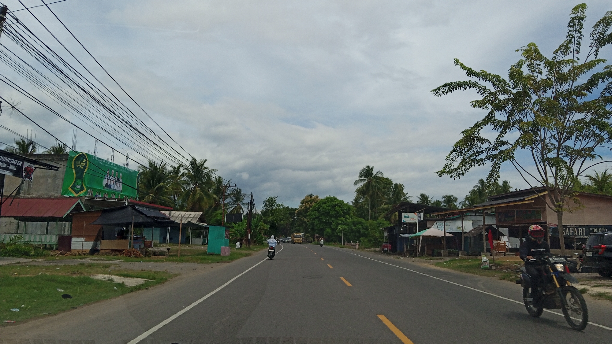 This is a medium-shot, eye-level, full-color photograph of a paved road in Kruengbaro, Indonesia, under a cloudy sky. The road runs horizontally across the frame, with two lanes of traffic visible. On the left side of the road, there are several small buildings, some with red roofs, and a large green banner with Arabic calligraphy and images. A motorcycle rider, wearing a helmet and dark clothing, is visible on the right side of the road. A car is parked on the right side of the road near a building with a sign that says "SAFARI". Tropical trees and vegetation are abundant throughout the scene. A yellow truck is seen in the distance on the road. Several power lines are strung across the top of the frame.