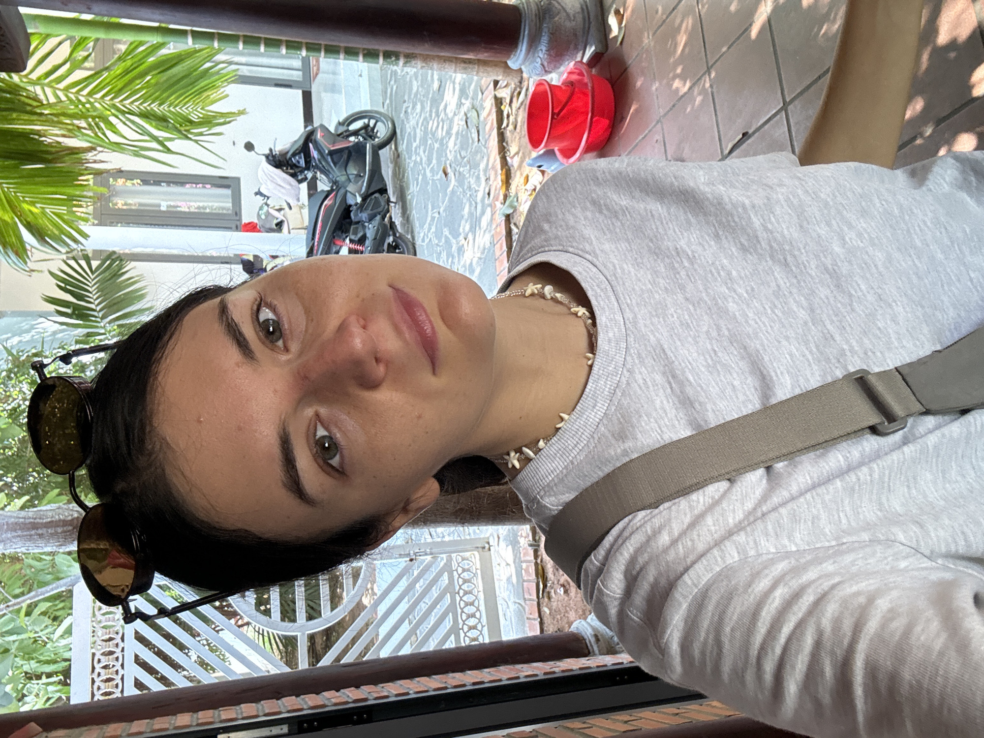 A young woman is posing for a selfie outdoors in Mũi Né, Vietnam. She is wearing a light gray t-shirt, a seashell necklace, and a tan strap across her body. Sunglasses are perched on her head. In the background, there is a parked motorcycle, a red bucket, palm trees, a white metal fence, and a structure made of dark bricks. The ground is tiled. The light suggests it is daytime.