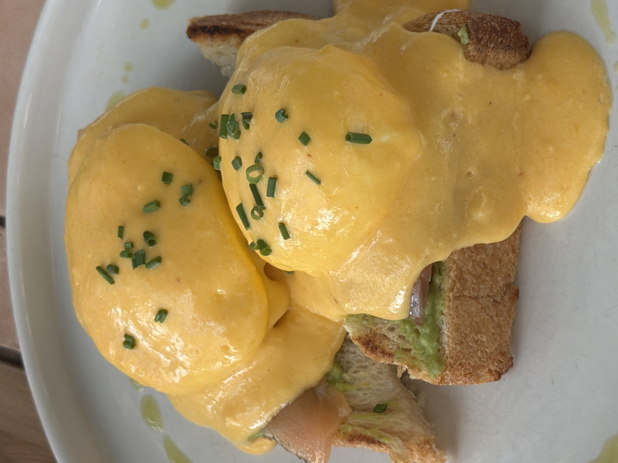 A close-up shot shows a serving of Eggs Benedict on a white plate. The dish consists of toast topped with avocado, smoked salmon, poached eggs, and hollandaise sauce, garnished with chopped chives. The setting appears to be a restaurant. The location and city details are not evident from the image.