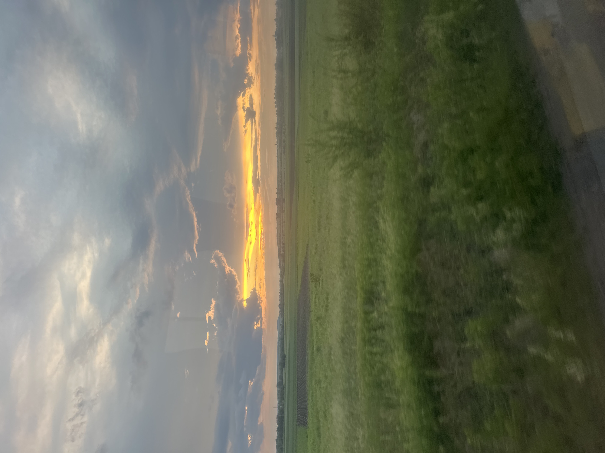 The image depicts a rural landscape, likely in or near Kulykiv, Ukraine, during sunset. The sky is filled with dramatic clouds, with a bright band of orange and yellow light illuminating the horizon where the sun is setting. Below the sky, a vast, green field stretches across the frame, appearing to be farmland with some sections showing tilled soil. In the foreground, the green vegetation is blurred, suggesting motion, perhaps captured from a moving vehicle. The overall scene evokes a sense of natural beauty and the end of the day.