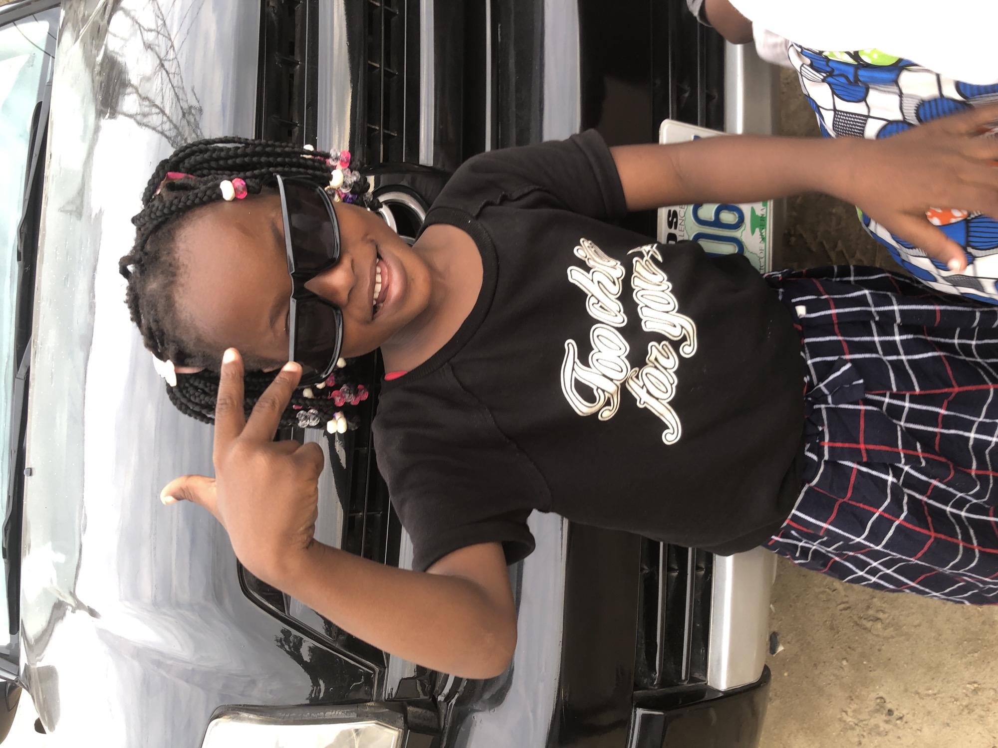 In Ado, Nigeria, a young girl, wearing black sunglasses and a black t-shirt with the text "Too chic for you" emblazoned in white, stands in front of a black vehicle. Her hair is styled in numerous braids adorned with colorful beads. She is playfully posing with her right hand raised, fingers forming a peace sign near her forehead. She is also wearing a patterned skirt. A license plate with the characters "OS 06" is visible on the front of the vehicle. The ground beneath her is composed of dry, sandy soil.