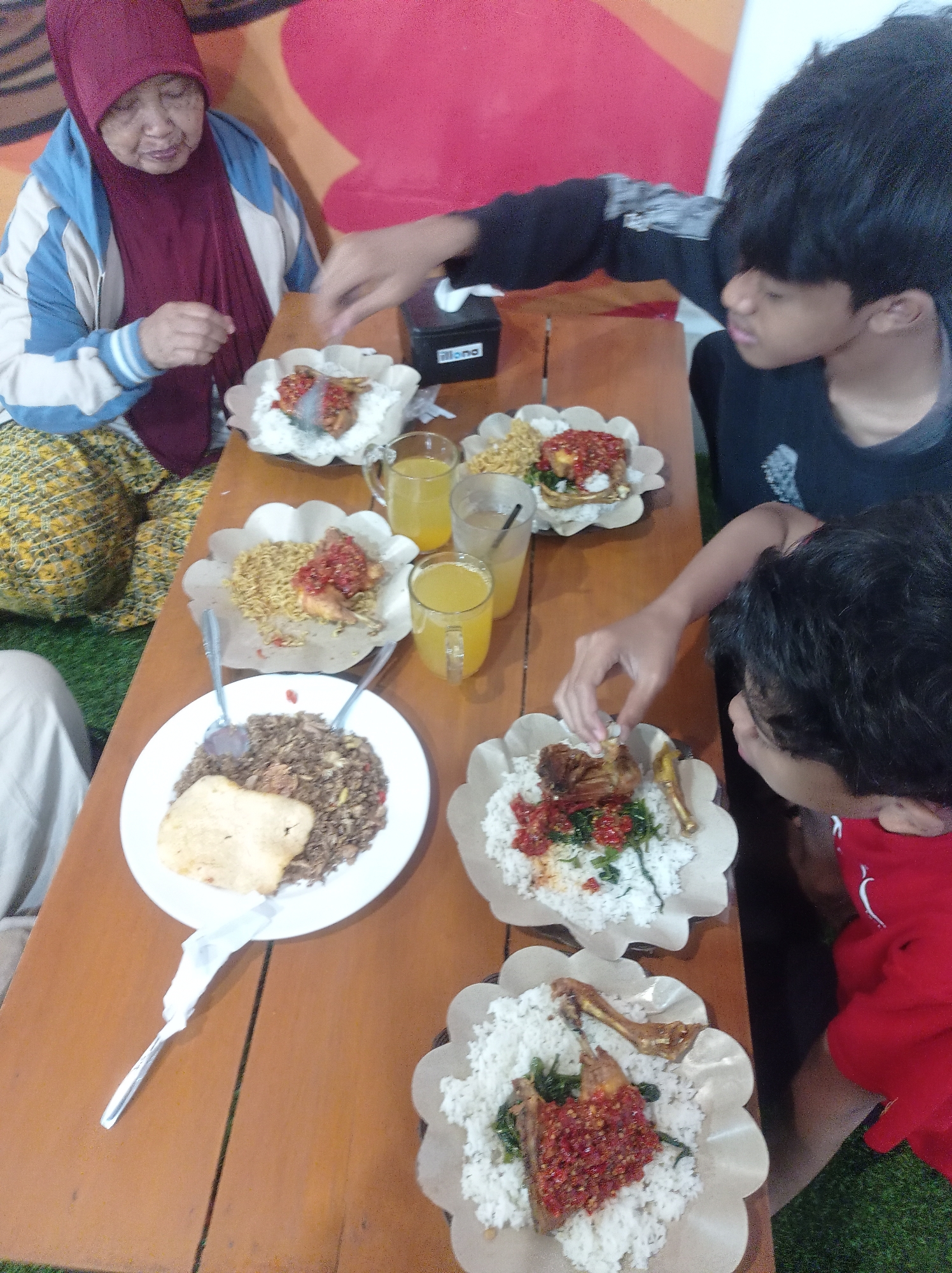 In Landungsari, Indonesia, a group of people are gathered around a wooden table, sharing a meal. An elderly woman, wearing a maroon hijab and a blue and white jacket, sits on the left. She is looking down at her food, with her right hand resting on the table. In front of her is a plate with white rice and what appears to be fried rice, topped with a light-colored cracker.
To the right, two young individuals are also eating. One, wearing a black t-shirt, is reaching for a plate of food that includes rice, fried chicken, and a vibrant red chili sauce. The other, in a red shirt, is actively eating from a similar plate, with a piece of fried chicken visible.
On the table are several plates of food, each containing rice, vegetables, and pieces of fried chicken or other protein, often adorned with a generous amount of chili sauce. Two glasses of orange juice are also present, along with a small black electronic device. The table is set on a green surface, possibly artificial turf, indicating an outdoor or semi-outdoor dining area. The background features a wall with abstract colorful patterns.