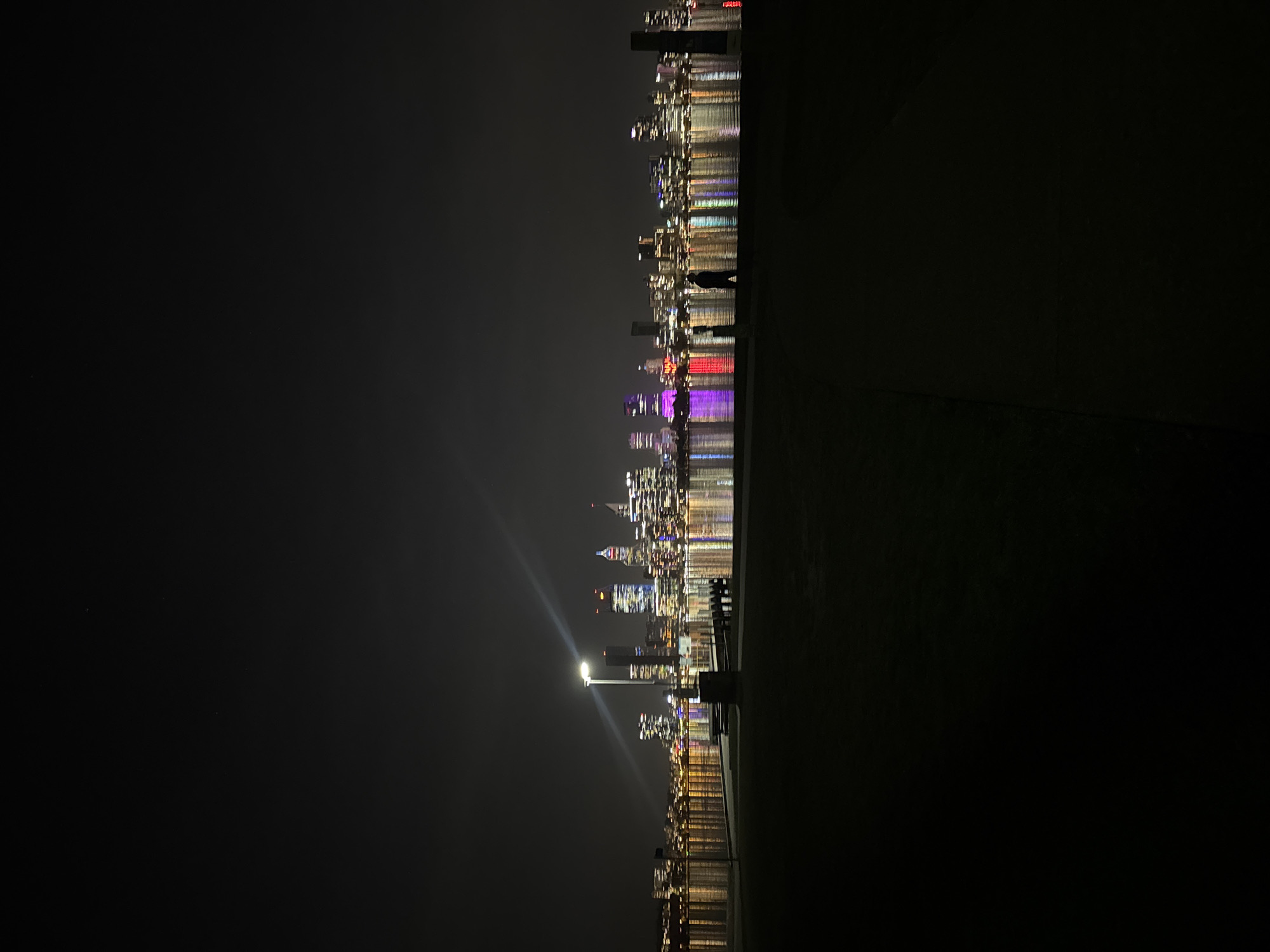 At night, the city skyline of South Perth, Australia, is reflected in the water. Buildings with colorful lights are visible across the dark water. A row of dark figures of people stand along the edge. A single streetlamp illuminates a bench and railing along the shore. The foreground is dominated by dark land.