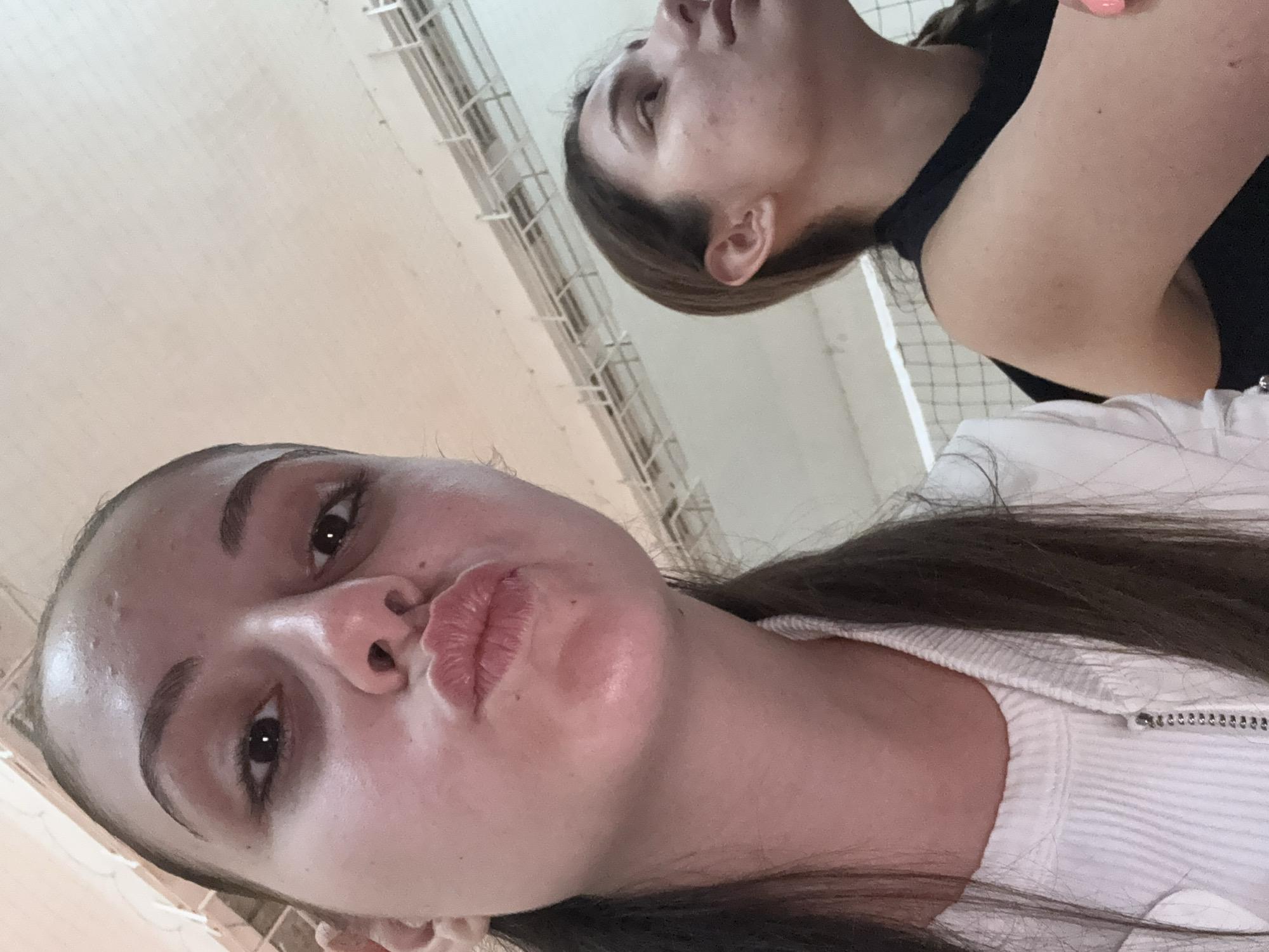 The image captures two young women indoors, likely in a facility in Burgas, Bulgaria, given the provided context. The perspective is a close-up, low-angle shot looking upwards.
In the foreground, a woman with dark hair, pulled back, looks directly at the camera with a playful "kissy" face. She has prominent eyebrows and dark eyes. She is wearing a white, ribbed top with a silver zipper visible on the collar. Her skin appears fair.
Behind her, partially out of focus, is another woman with light brown hair, seen in profile. She is looking upwards and to the right of the frame, her expression appearing serene or contemplative. She wears a black top, and a hand with pink nail polish is visible near her shoulder.
The background features light-colored walls or a ceiling with visible white metal grilles or netting, suggesting an indoor sports facility, gymnasium, or a similar public recreational building. The lighting is uniform, indicative of artificial indoor illumination. No text is visible in the image. The overall atmosphere is casual and familiar.