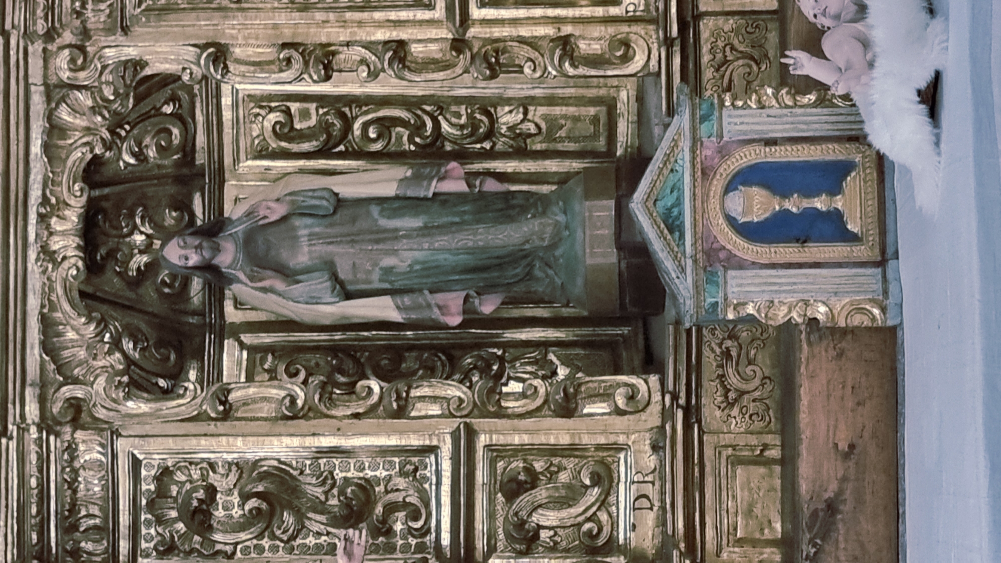 A vertical, medium shot captures a religious altar with a statue of Jesus Christ as the central figure, positioned behind a small, ornate shrine. The altar's backdrop is an elaborate gilded wooden structure, featuring carved scrollwork and paneling. To the left of Jesus, a hand, possibly part of another statue or a viewer's interaction with the altar, is visible, partially obscured by the gilded relief. The statue of Jesus is depicted with a serene expression, long hair, and is draped in a green robe with pink trim. His hands are raised, with fingers extended, in a gesture of blessing or teaching. Below Jesus, a small shrine or tabernacle is adorned with a gold chalice against a blue background, framed by columns and decorative molding. To the far right of the frame, a small, plump infant figure, appearing to be a statue of baby Jesus, lies on a fluffy white material. The infant's right arm is raised, and its face is turned towards the viewer. The altar is situated on a flat, light-colored surface, possibly a shelf or a pew, that extends into the foreground. The overall impression is one of a richly decorated, devotional space within a church or chapel.
