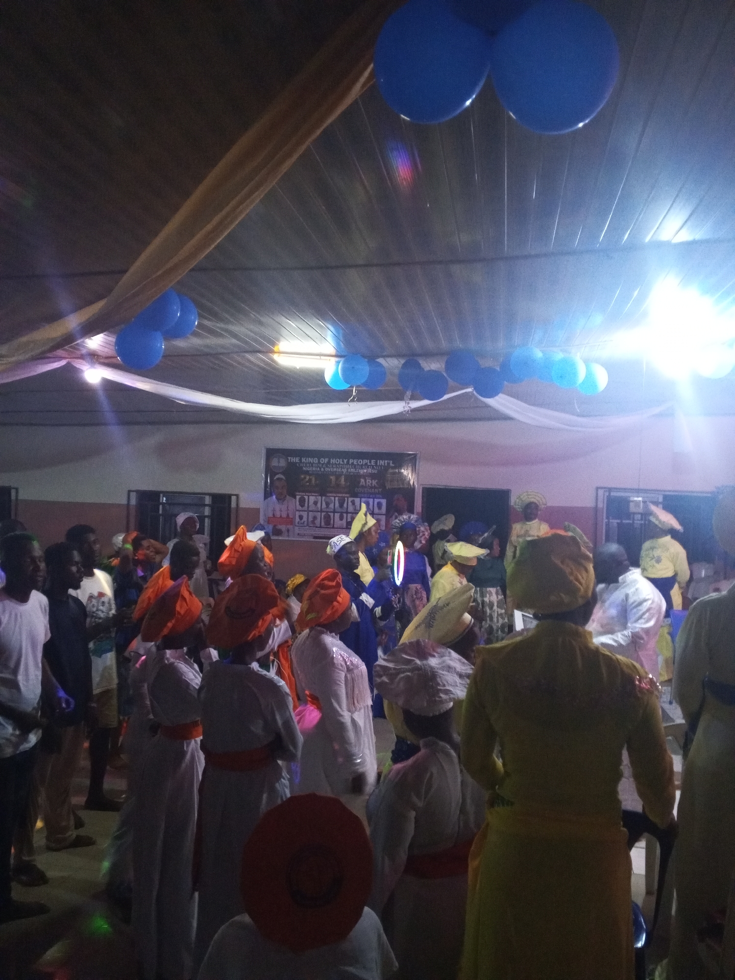 People are gathered indoors in what appears to be a hall decorated with blue balloons and draped fabric. A banner or poster is visible in the background with text including "THE KING OF HOLY PEOPLE INT'L" and "ABK." The gathering includes individuals wearing traditional attire, such as orange head wraps and white robes with orange sashes. Other people are dressed casually in plain clothing. The lighting is bright and appears to be from artificial sources. Location cannot be confirmed as Lokoja, Nigeria.