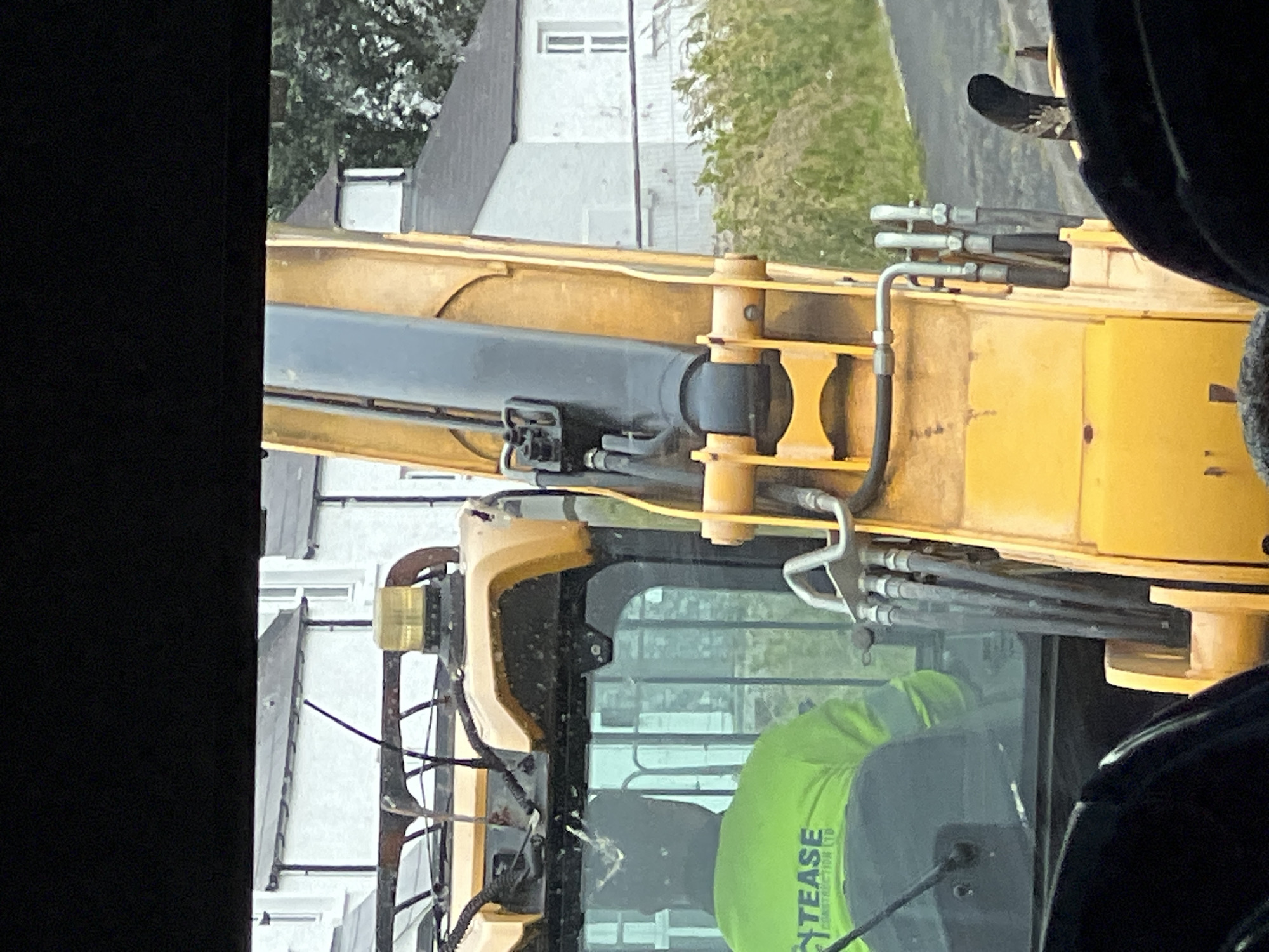 The image captures a close-up view of a yellow excavator. The operator, wearing a bright green high-visibility jacket with the text "TEASE CONSTRUCTION LTD" visible, is seated inside the cab. The excavator's arm is prominently featured, with its hydraulic hoses and mechanisms clearly visible. In the background, a row of white houses with dark roofs suggests a residential area. The presence of an excavator implies construction or maintenance work is underway. The overall setting appears to be outdoors, likely in a rural or semi-rural part of Portnablagh, Ireland, given the architectural style of the houses and the surrounding vegetation. The lighting suggests it is daytime.
