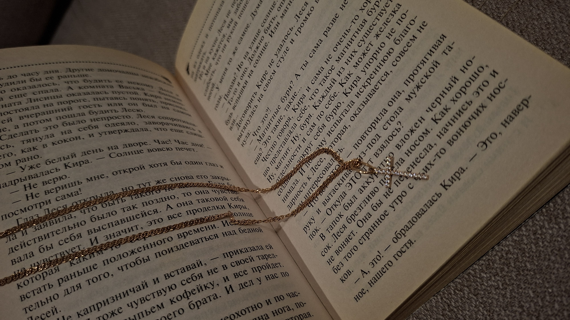 A medium shot depicts an open book with Cyrillic text. A gold-colored chain necklace with a cross pendant rests atop the pages. The book's paper is aged, with a yellow hue. The text appears dense and closely spaced. The chain is finely linked. The pendant, a cross, is adorned with small, possibly rhinestone, details. The book lies on a soft, possibly upholstered, surface with a neutral tone.