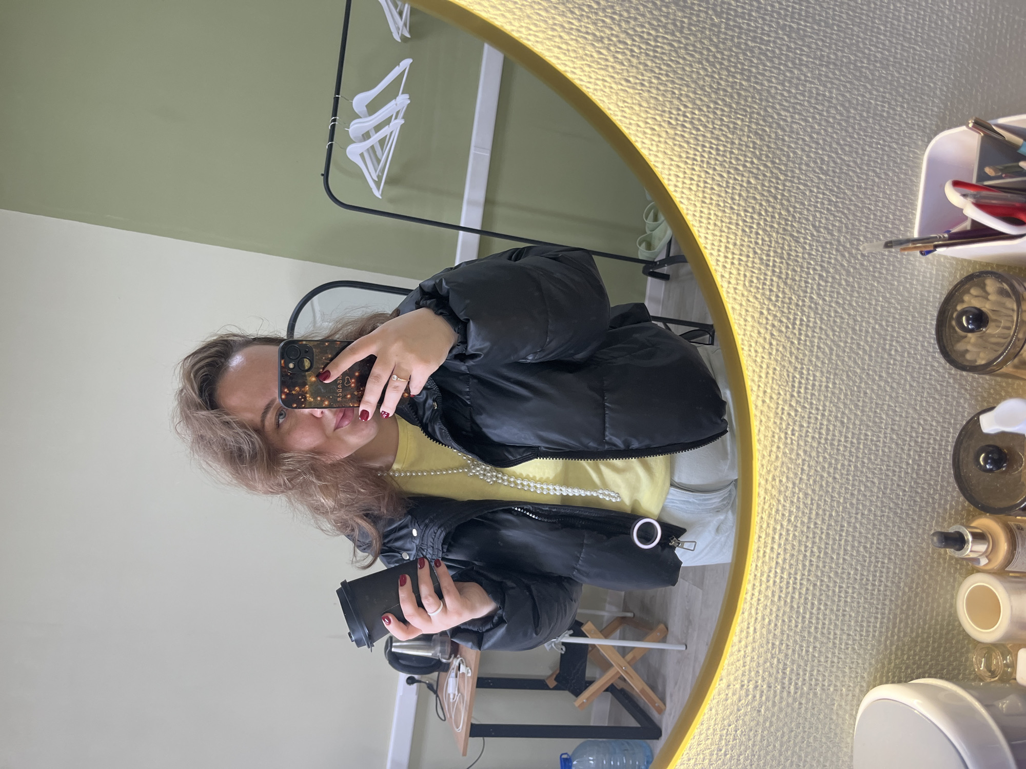 A woman is captured taking a mirror selfie indoors. She has light brown, wavy hair and is dressed in a black puffer jacket over a light yellow top, adorned with a long pearl necklace. Her left hand, with red-painted nails and a ring, holds a smartphone featuring a dark, patterned case with indistinct reversed text. In her right hand, she holds a black disposable coffee cup. She looks towards the camera, reflecting her face and upper body in the mirror.
The setting is an indoor space, likely a dressing room or salon, suggested by the surrounding elements. The walls reflected in the mirror are painted in two tones: light green on the upper section and a lighter beige below. A black metal clothing rack with several white hangers is visible in the reflection.
Outside the mirror's frame, to the right, a collection of items is arranged: a white container filled with tools like scissors and brushes, a clear jar of cotton buds, and various cosmetic bottles including one with a dropper and another with a pump dispenser. Below the mirror, a wooden folding stool, a blue water bottle, and some white charging cables are visible on the light-colored floor. The round mirror has an illuminated, textured golden frame, emitting a warm light that suggests a well-lit interior, likely during daytime. The location is in Chaikovsky, Russia.