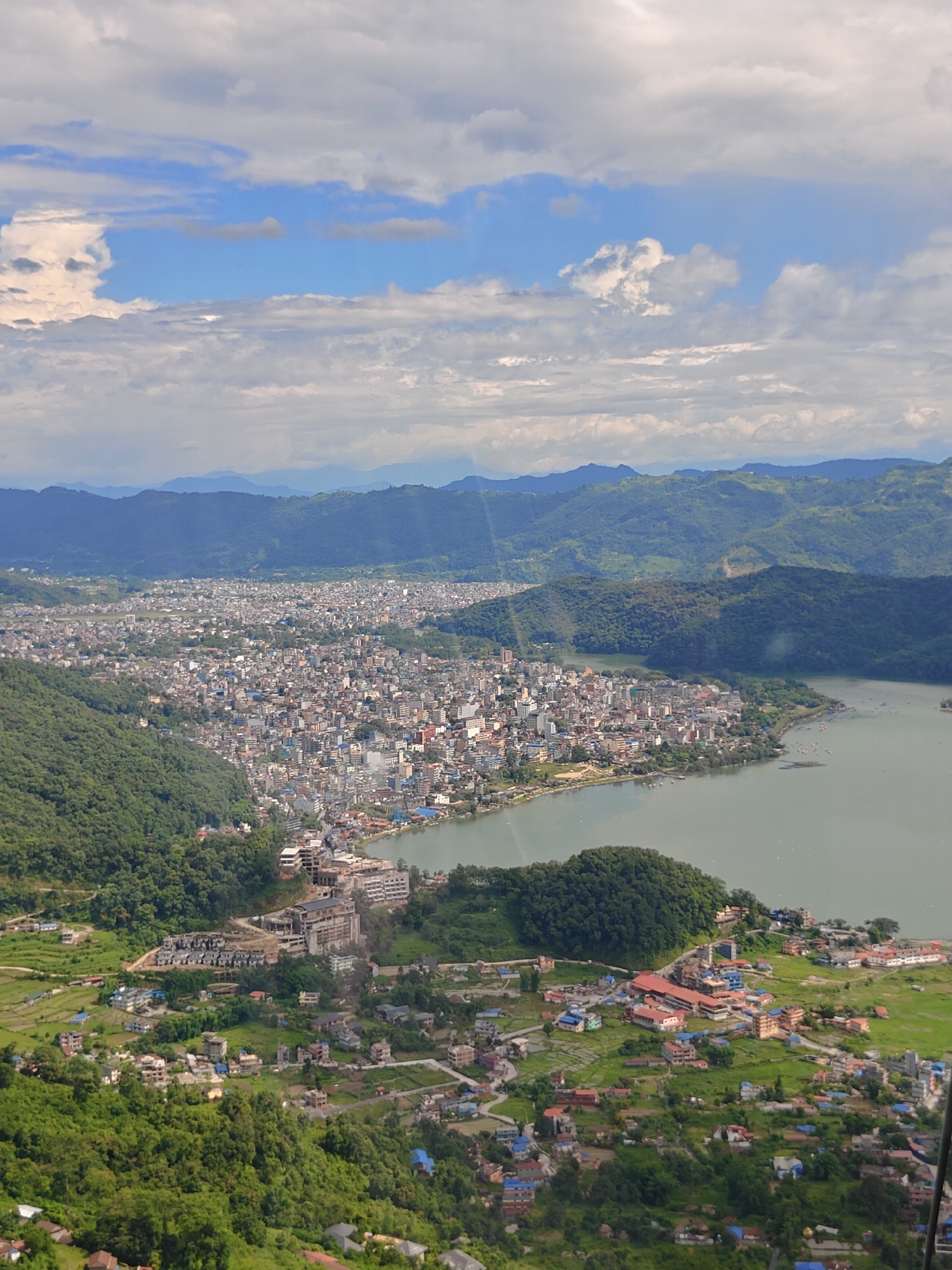 This is an aerial view of the city of Pokhara, Nepal, looking east over Phewa Lake and the surrounding valley. The dense urban area of Pokhara stretches along the northern edge of the lake, with buildings densely packed together. The lake itself is a calm, greenish-blue body of water, with a few small boats visible on its surface.
In the foreground and to the left, the landscape is dominated by lush green hillsides covered in dense vegetation. These hills are interspersed with smaller pockets of development, including houses, agricultural fields, and some construction sites with visible building materials. Towards the lower right of the frame, there are more scattered houses and buildings with red tiled roofs.
Further in the distance, a series of rolling hills and mountains form a backdrop to the city, with the shadows of clouds casting patterns across their slopes. The sky is a mix of blue with scattered white and grey clouds, some of which are large and puffy, indicating a dynamic weather system. The overall impression is of a vibrant, populated city nestled within a beautiful, mountainous natural environment.