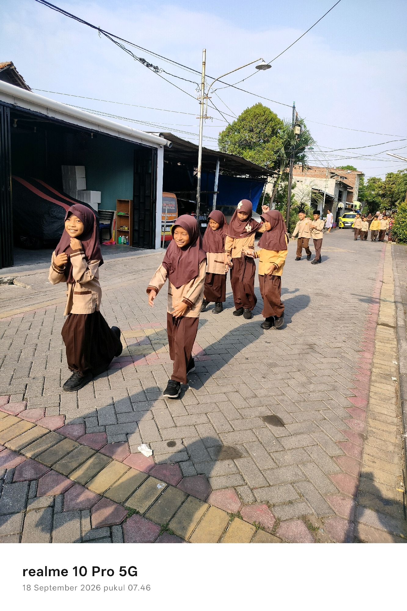 The image depicts a group of young schoolchildren, presumably on their way to or from school, walking along a paved sidewalk in what appears to be a residential area in Semarang, Indonesia. The children are all wearing similar uniforms consisting of light brown tops with darker brown accents and dark brown headscarves or caps, and dark brown skirts or trousers. They are walking in a line, with some children in the foreground more clearly visible and others further in the background.
In the foreground, two girls are prominently featured. The girl on the left is smiling and adjusting her headscarf, while the girl on the right is also smiling and looking forward as she walks. Their attire suggests a religious school uniform, common in Indonesia.
The environment consists of a paved walkway with a patterned brick design in shades of grey, yellow, and pink. Shadows cast by the children and overhead utility poles are visible on the ground. To the left of the walkway, there is a building with a white roof and dark-colored walls, possibly a shop or a house with an open garage. In the background, more buildings, trees, and utility poles are visible, indicative of an urban or suburban setting. A yellow vehicle, possibly a car or small bus, is seen further down the street, along with other people walking. The sky is bright blue, suggesting a sunny day. The text at the bottom of the image indicates the date and time the photo was taken: "18 September 2026 pukul 07.46".