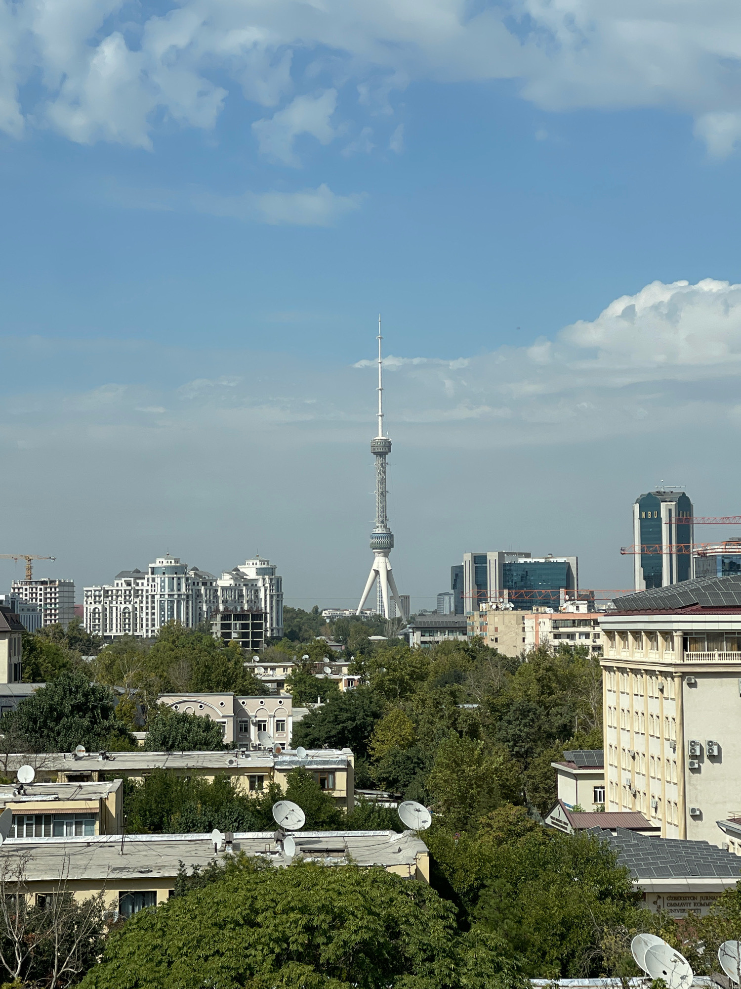 The image captures a panoramic view of the city of Tashkent, Uzbekistan, dominated by the iconic Tashkent Television Tower, a prominent landmark. The tower stands tall in the mid-ground, its lattice structure reaching towards a partially cloudy blue sky. Surrounding the tower are various buildings, including modern high-rises with glass facades and more traditional residential buildings with lighter colored exteriors. Lush green trees are interspersed throughout the cityscape, particularly in the foreground and mid-ground, adding a natural element to the urban environment. Several satellite dishes are visible on the rooftops of some of the lower buildings. Construction cranes are also present in the distance, indicating ongoing development in the city. The overall scene suggests a bright, daytime view of a bustling metropolis.