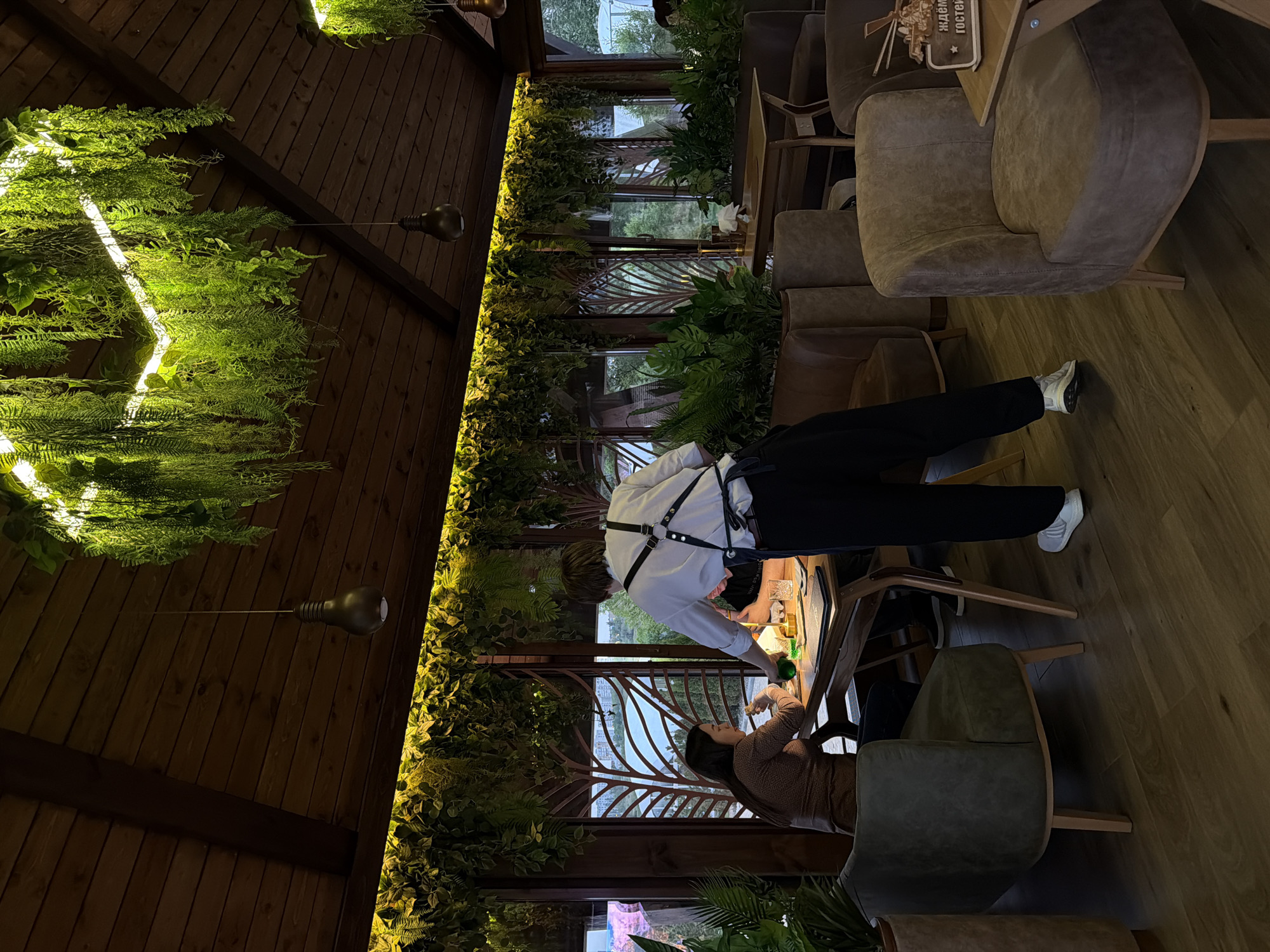 The image depicts the interior of a restaurant or cafe. The space features a wooden ceiling and lush greenery, with plants hanging from the ceiling and adorning the walls. There is a person in a white shirt and dark pants standing at a table, appearing to interact with another person seated at the table. The other individual is seated and wearing a brown sweater. The cafe features stylish chairs, and natural light streams in through the windows. There is an individual serving the seated person. The location is Kamyshin, Russia.