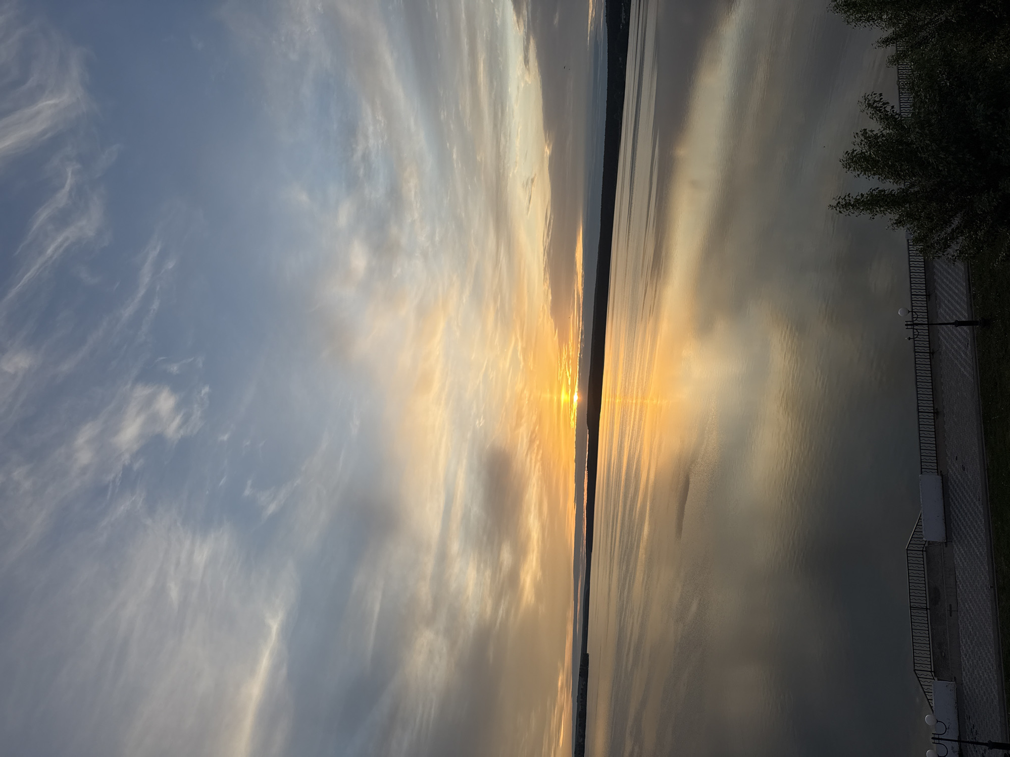 The image depicts a sunset over a body of water, likely the Votkinsk Reservoir, near Votkinsk, Russia. The sky is filled with clouds illuminated by the setting sun, casting a golden glow on the water's surface. A dark strip of land is visible in the distance, likely the opposite shore of the reservoir. In the foreground, a paved walkway with a railing and lampposts is present, alongside some greenery. The image is rotated 90 degrees counterclockwise.